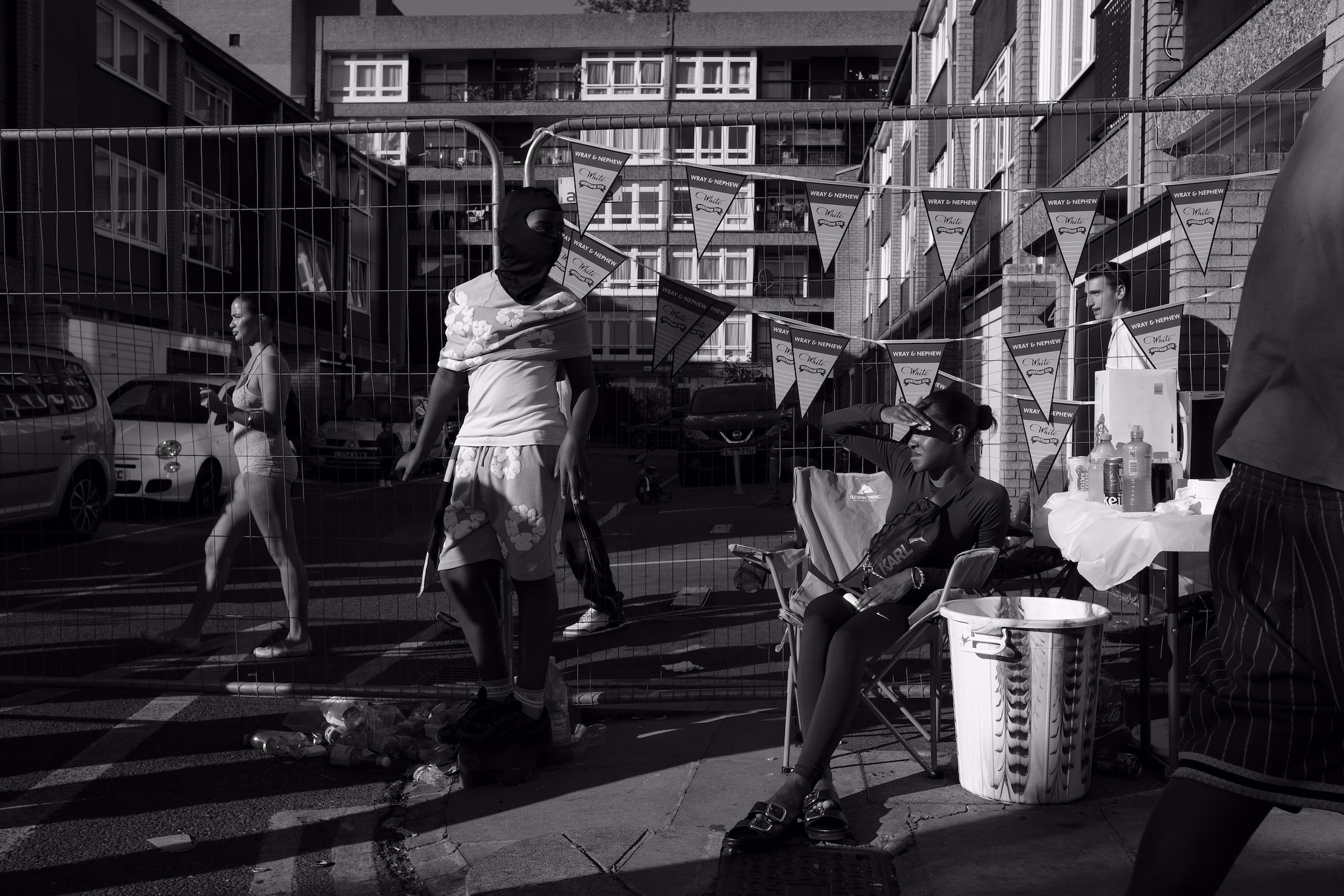 Des personnes dans une rue urbaine décorée de fanions, avec un mur grillagé et des bâtiments en arrière-plan.