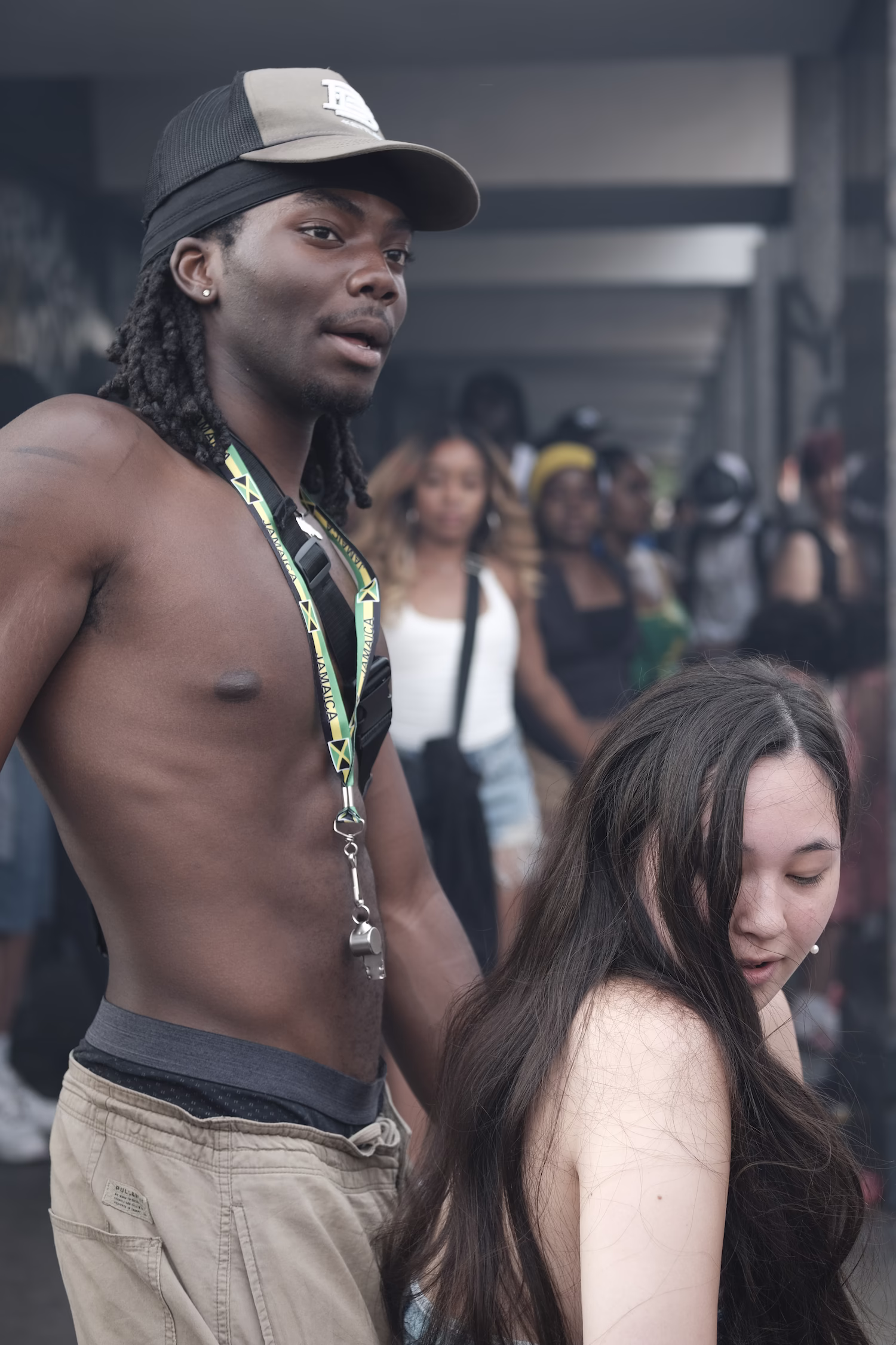 Homme torse nu avec casquette et lanière Jamaïque, femme aux longs cheveux bruns devant lui en foule floue.