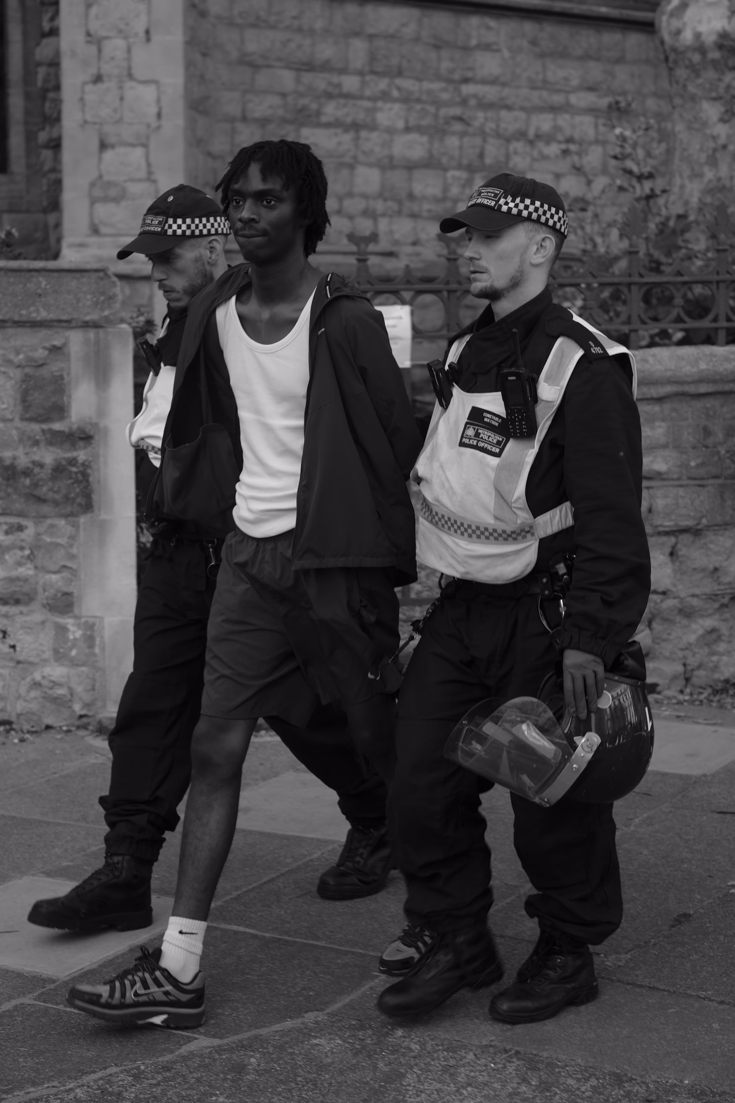 Un homme menotté encadré par deux policiers en uniforme marchant devant un mur en pierre.
