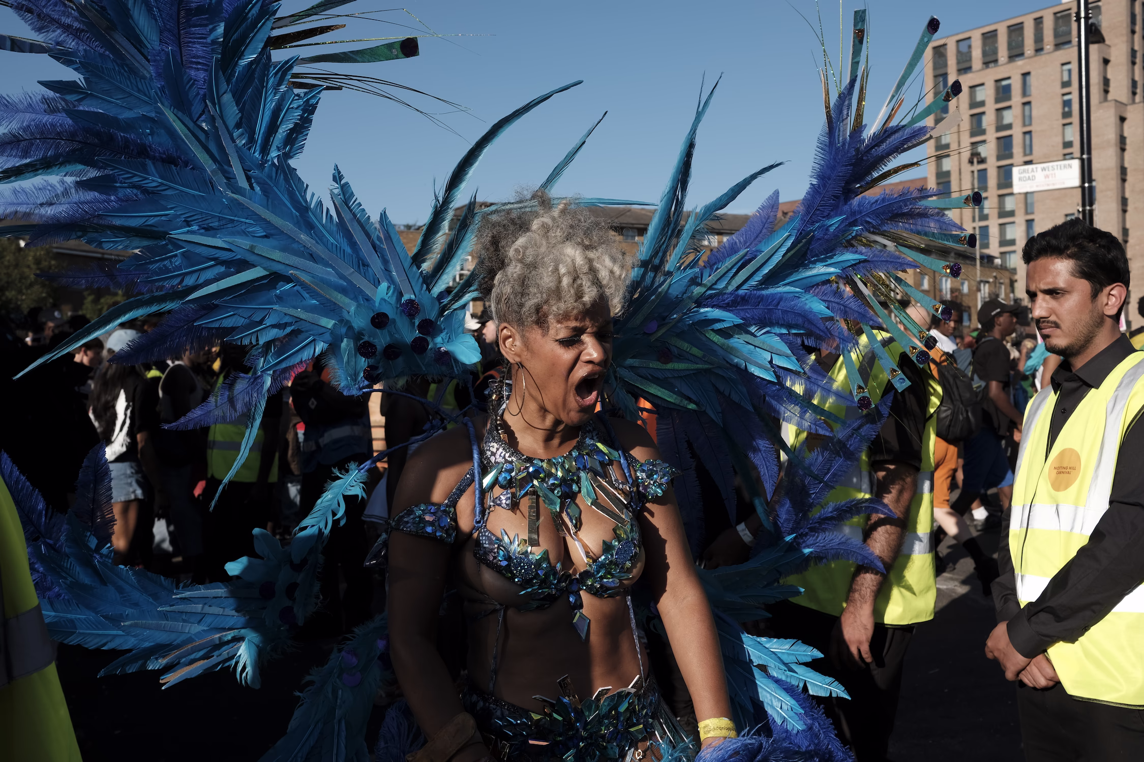 Femme en costume bleu orné de plumes lors d'un carnaval, avec des spectateurs en arrière-plan.