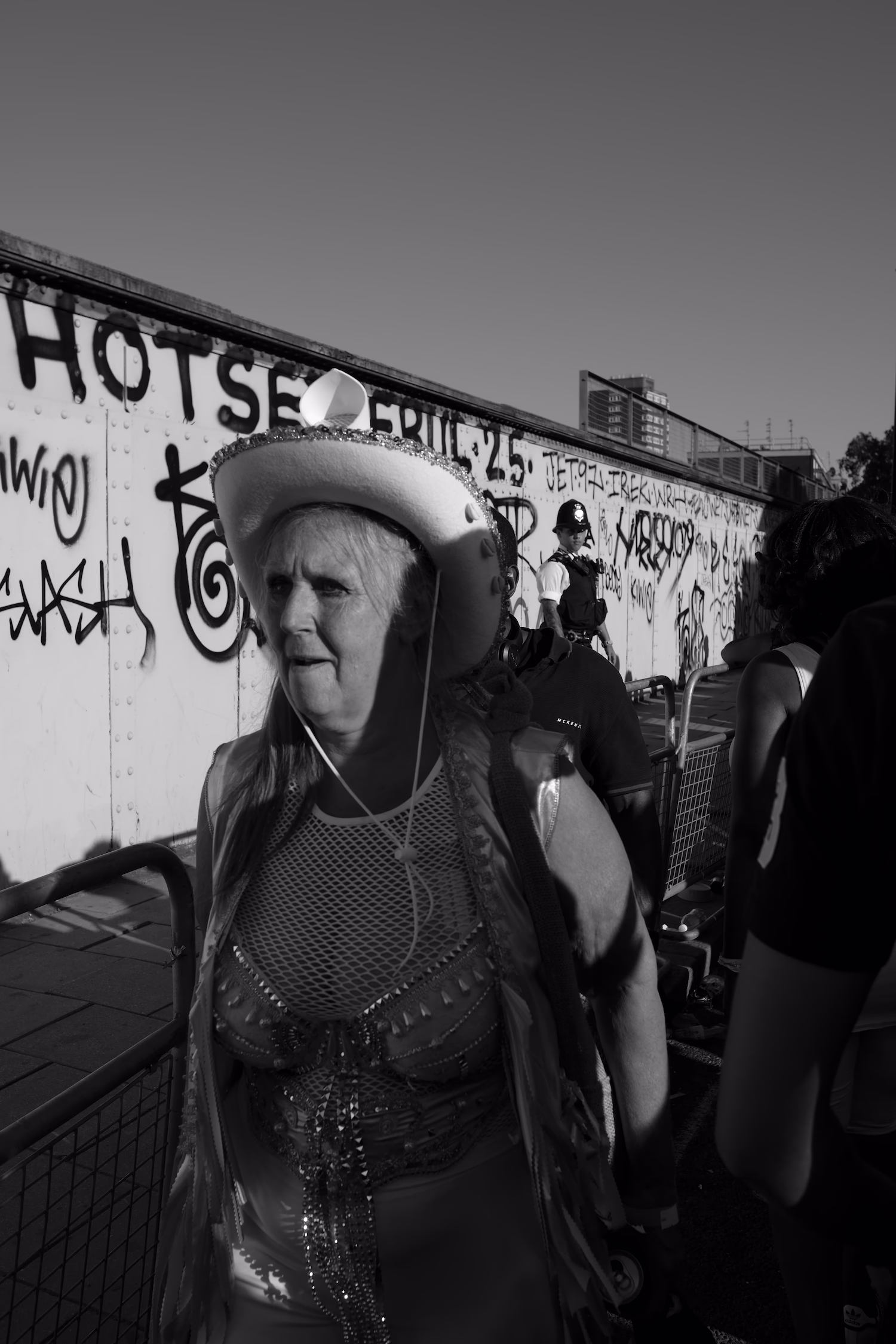 Femme en costume festif et chapeau marche devant un mur tagué avec un policier en arrière-plan.