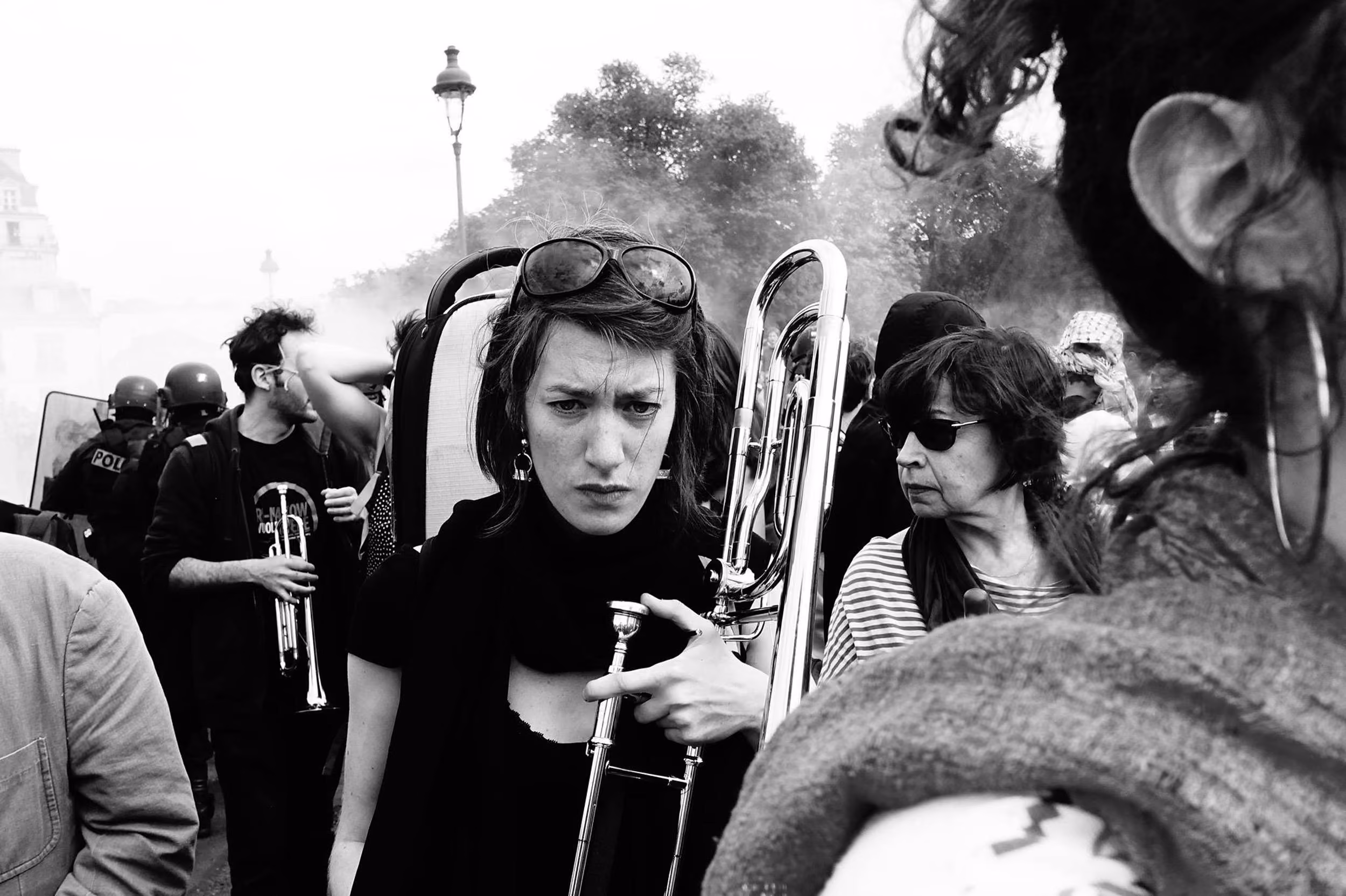 Manifestation en extérieur avec plusieurs personnes, dont une femme au premier plan portant des lunettes de soleil sur la tête et tenant un trombone, avec une expression sérieuse, entourée d'autres participants et de policiers en arrière-plan.