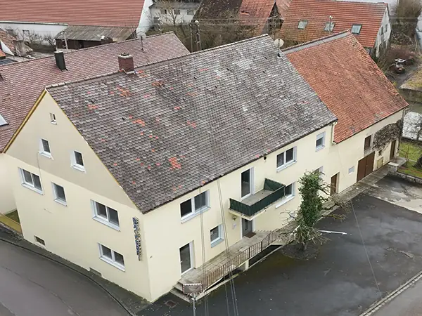 Luftaufnahme eines beigefarbenen zweistöckigen Gebäudes mit Ziegeldach, das einige Abnutzungserscheinungen aufweist und an einer Straßenecke in einer Kleinstadt steht. Das Gebäude hat einen kleinen Balkon, und der Bereich um das Gebäude ist gepflastert.