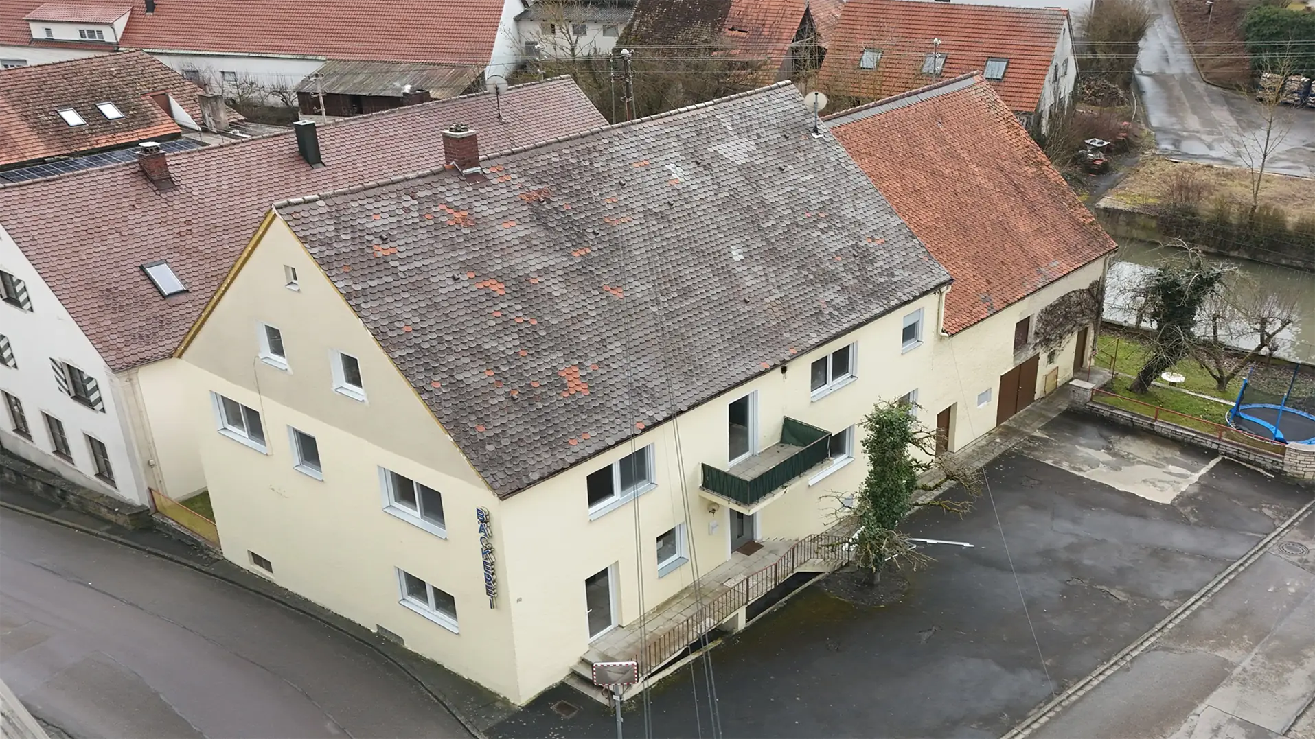 Luftaufnahme eines beigefarbenen zweistöckigen Gebäudes mit Ziegeldach, das einige Abnutzungserscheinungen aufweist und an einer Straßenecke in einer Kleinstadt steht. Das Gebäude hat einen kleinen Balkon, und der Bereich um das Gebäude ist gepflastert.