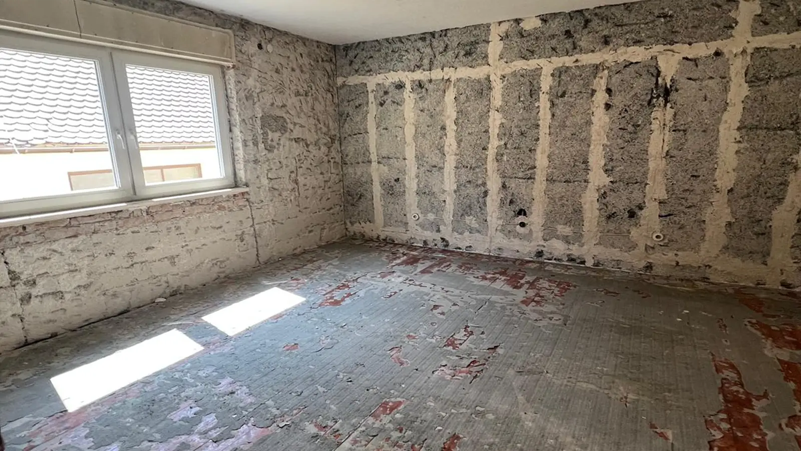 Ein leerer, ausgeräumter Raum mit kahlen, beschädigten Böden und unfertigen, freiliegenden Wänden. Durch ein großes Fenster dringt Sonnenlicht ein und erhellt das raue, renovierte Innere. Es sind weder Möbel noch Dekoration vorhanden.