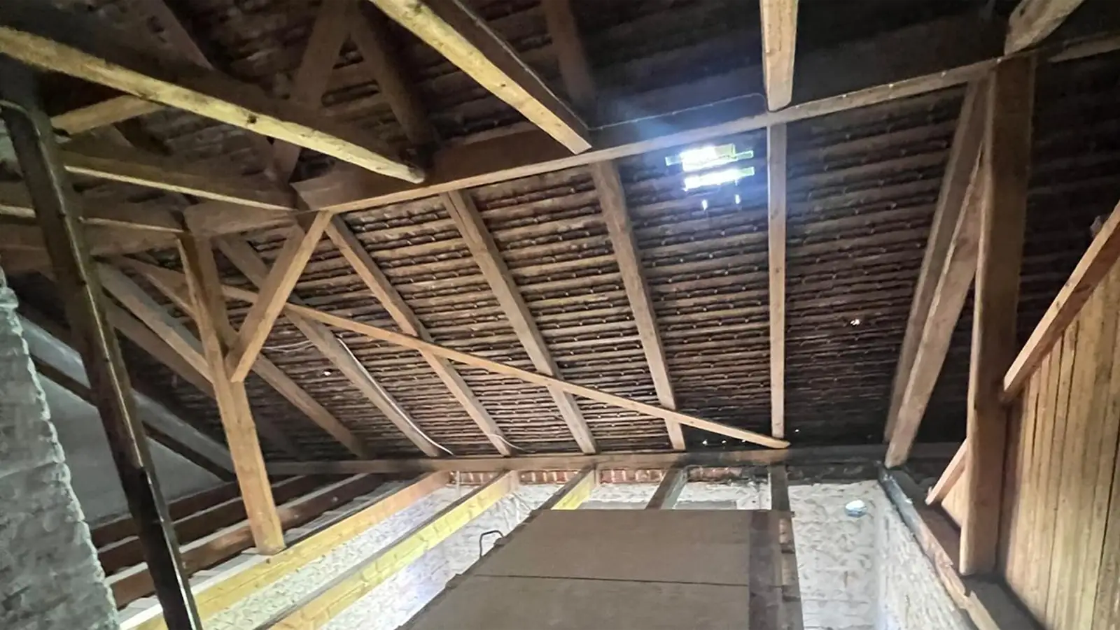 Blick auf einen unausgebauten Dachboden mit freiliegenden Holzbalken, Dachstützen und einem kleinen Lichtfleck, der durch eine Lücke im Dach fällt. Darunter sind einige Sperrholzplatten und Dämmstoffe zu sehen.