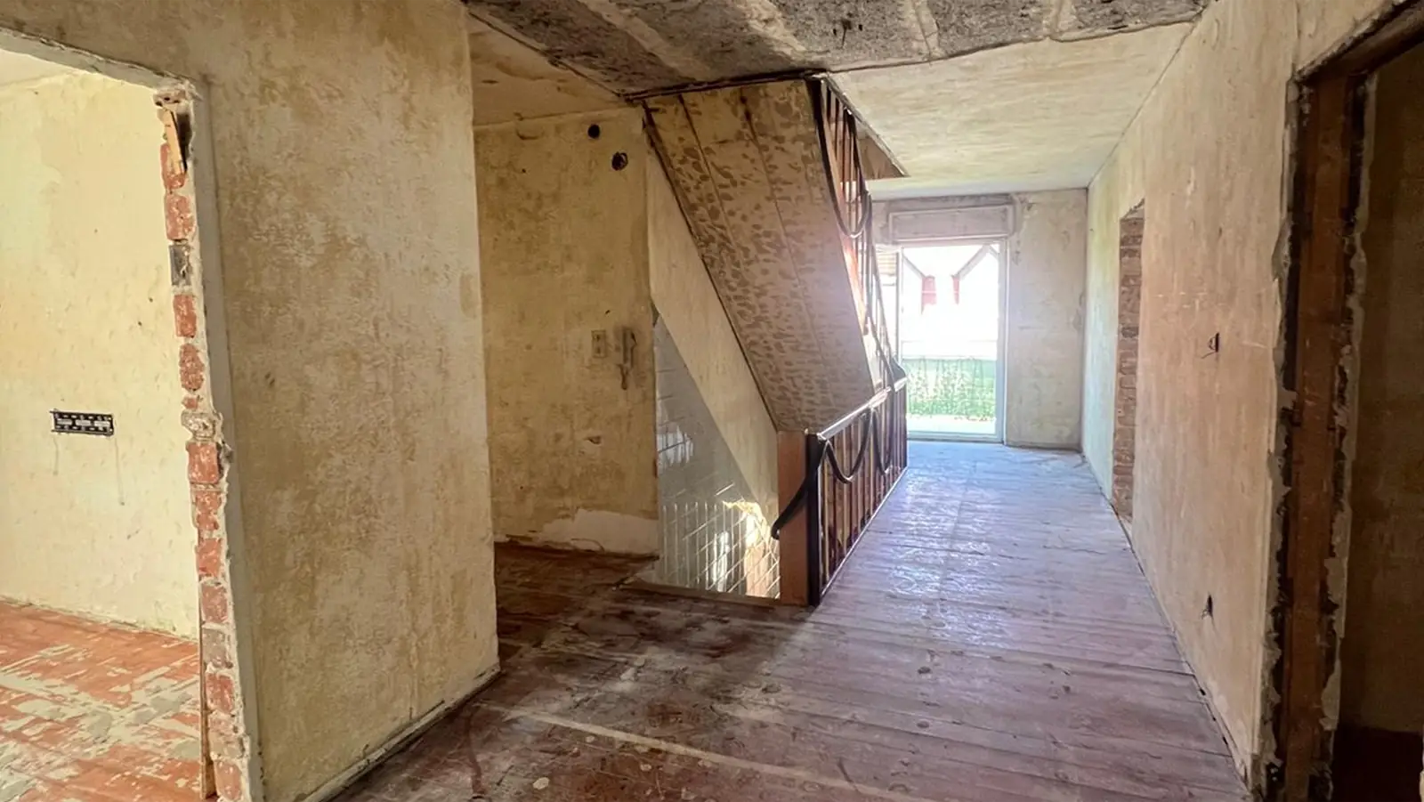 Flur eines unfertigen Gebäudes mit abgenutzten Böden, freigelegten Ziegeln an den Türrahmen und einer Treppe mit Metallgeländer. Das Sonnenlicht scheint durch eine offene Tür am Ende und gibt einen Blick ins Freie frei.