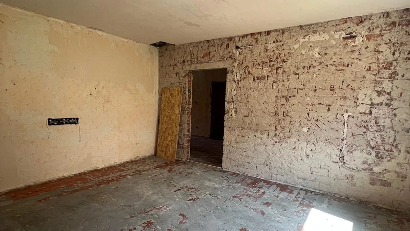 Ein leerer Raum mit freiliegenden Ziegelwänden, einem rauen Betonboden und einer unfertigen Türöffnung. Ein Stück Holz lehnt an einer Wand, und es sind weder Möbel noch Dekoration zu sehen.