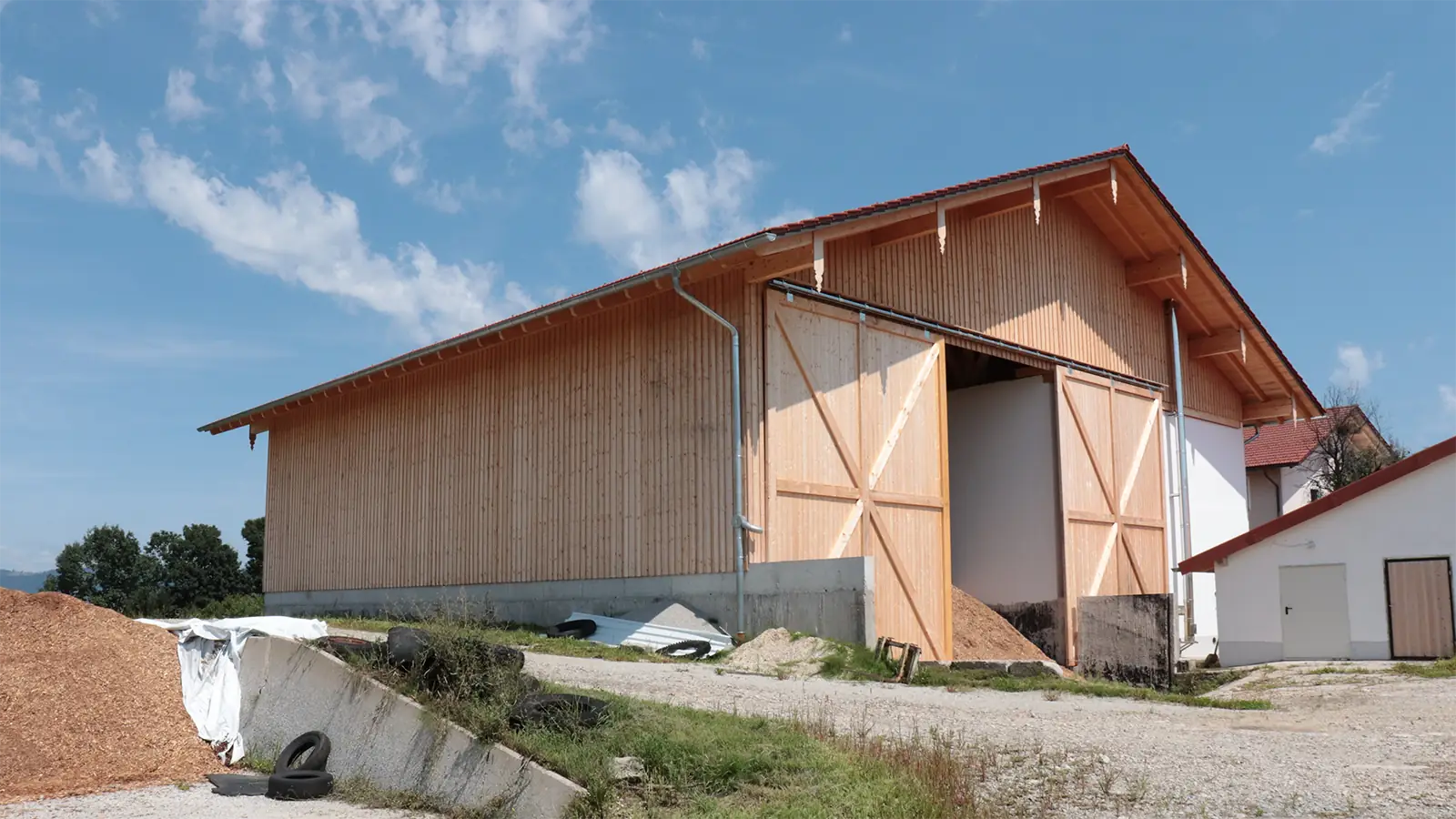 Eine große Lagerhalle mit Holzfassade und rotem Dach und weit geöffneten Türen steht auf einem Betonfundament, umgeben von einem Haufen Holzspäne und einigen verstreuten Autoreifen unter einem teilweise bewölkten, blauen Himmel.