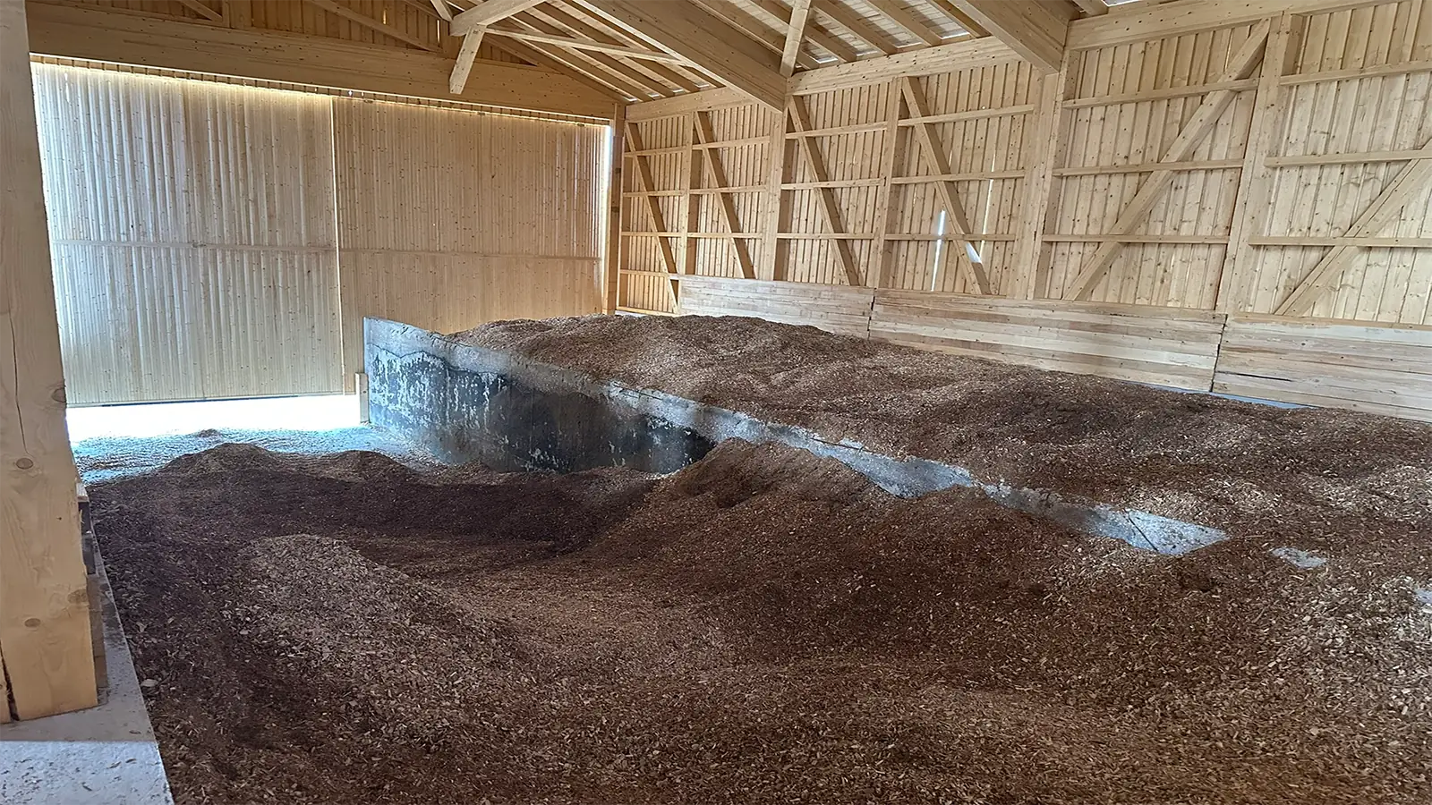 Ein großer Haufen Holzspäne oder Rindenmulch füllt einen Teil einer Lagerhalle aus Holz mit offenen Dachsparren und natürlichem Licht, das durch die Lücken in den Holzwänden scheint.