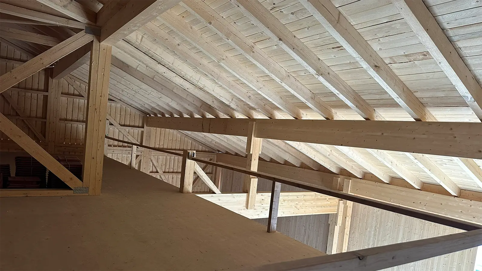 Innenansicht eines Dachbodens aus Holz mit freiliegenden Balken und Dachsparren, hellem Tageslicht und einem schmalen Gang mit einem Holzgeländer an einer Seite.