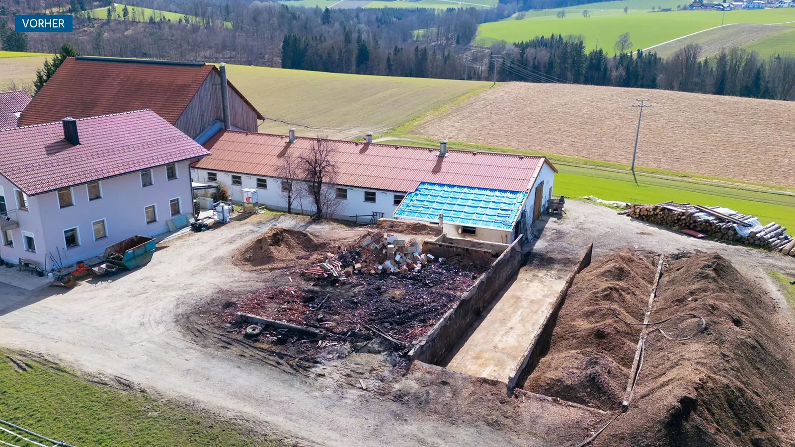 Luftaufnahme eines Hofs mit mehreren Gebäuden, Erd- oder Mulchhaufen, Trümmern auf einer Freifläche und umliegenden Feldern und Bäumen unter klarem Himmel.