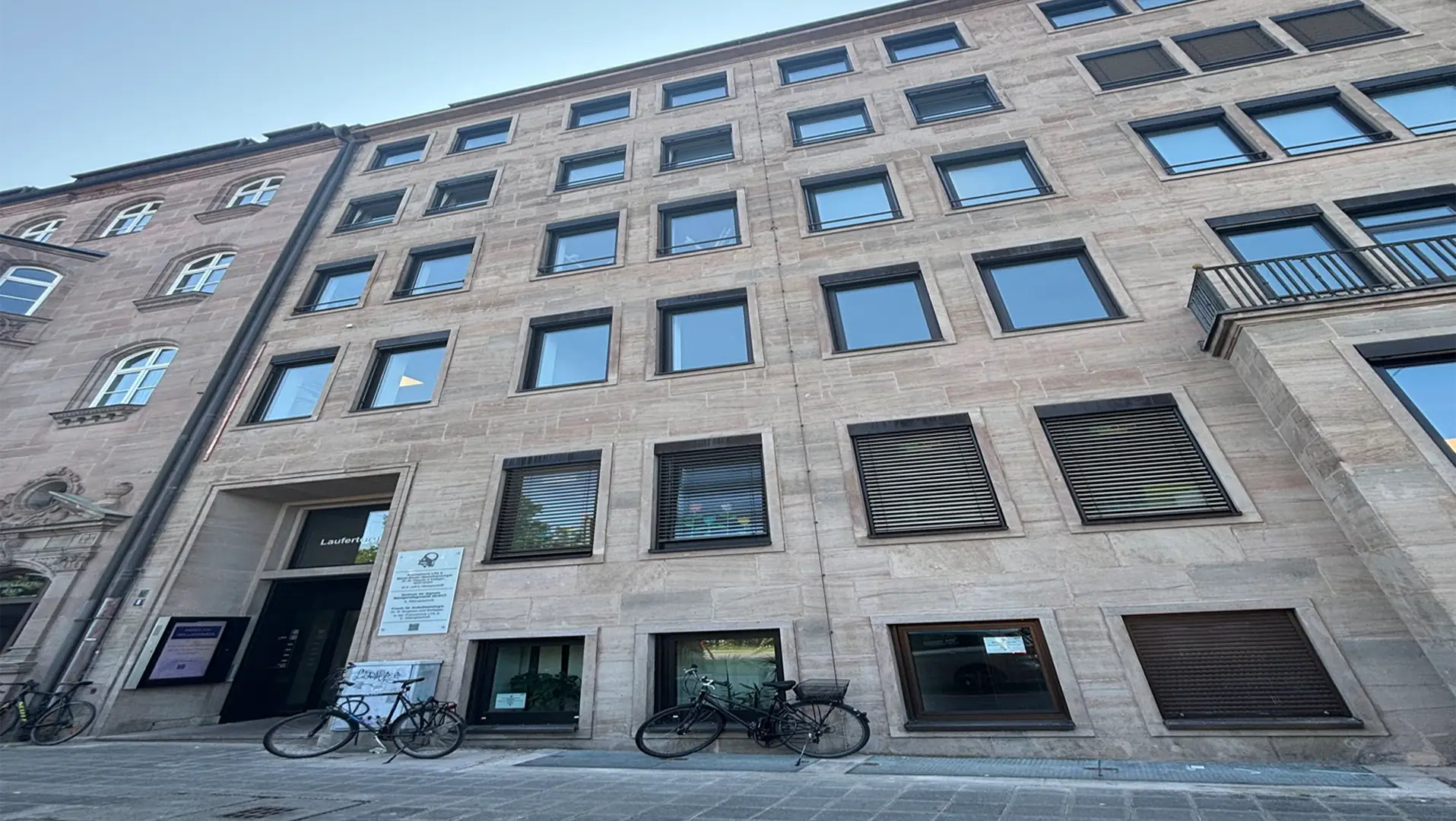 Ein hohes, beigefarbenes Steingebäude mit vielen rechteckigen Fenstern, teilweise mit Fensterläden, steht an einer gepflasterten Straße. Davor sind mehrere Fahrräder geparkt, und in der Nähe des Eingangs sind Schilder zu sehen. Über uns ist der Himmel klar