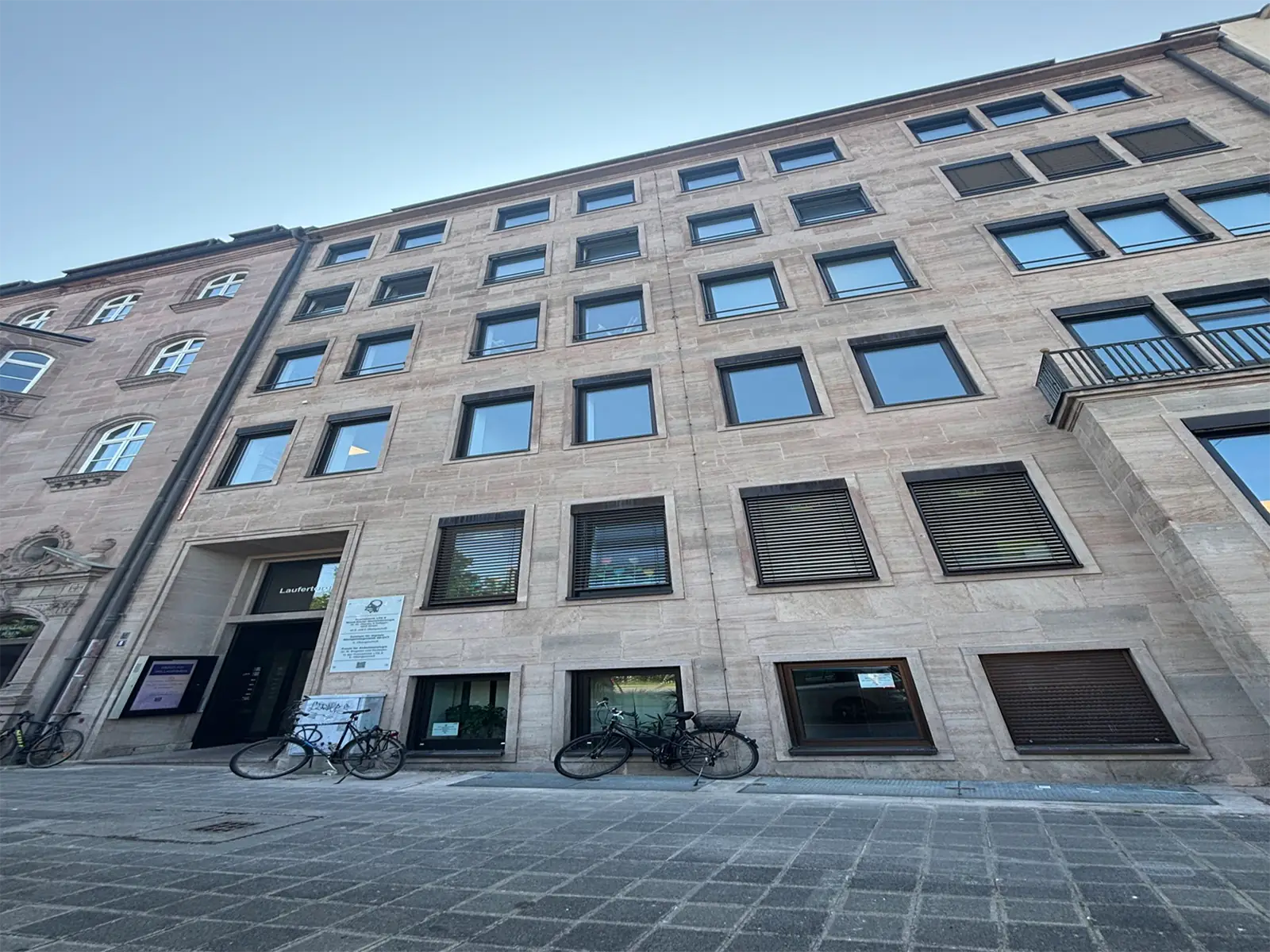 Ein hohes, beigefarbenes Steingebäude mit vielen rechteckigen Fenstern, teilweise mit Fensterläden, steht an einer gepflasterten Straße. Davor sind mehrere Fahrräder geparkt, und in der Nähe des Eingangs sind Schilder zu sehen. Über uns ist der Himmel klar