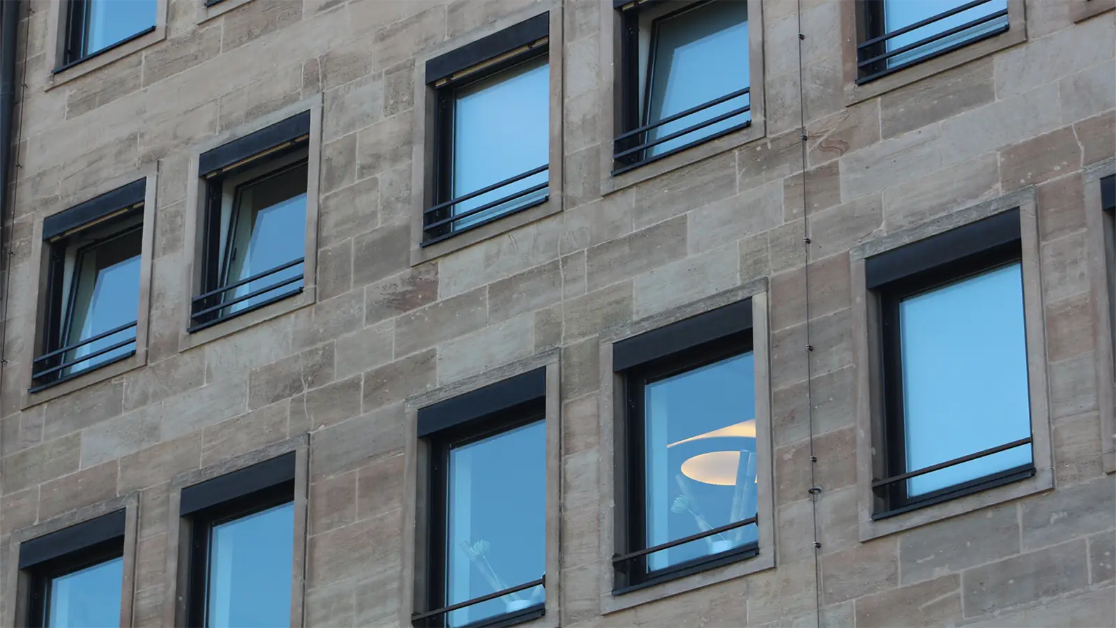 Nahaufnahme der Fassade eines Gebäudes mit mehreren rechteckigen Fenstern in einer Stein- oder Betonwand. In einigen Fenstern spiegelt sich der Himmel, während eines eine Innenbeleuchtung zeigt.