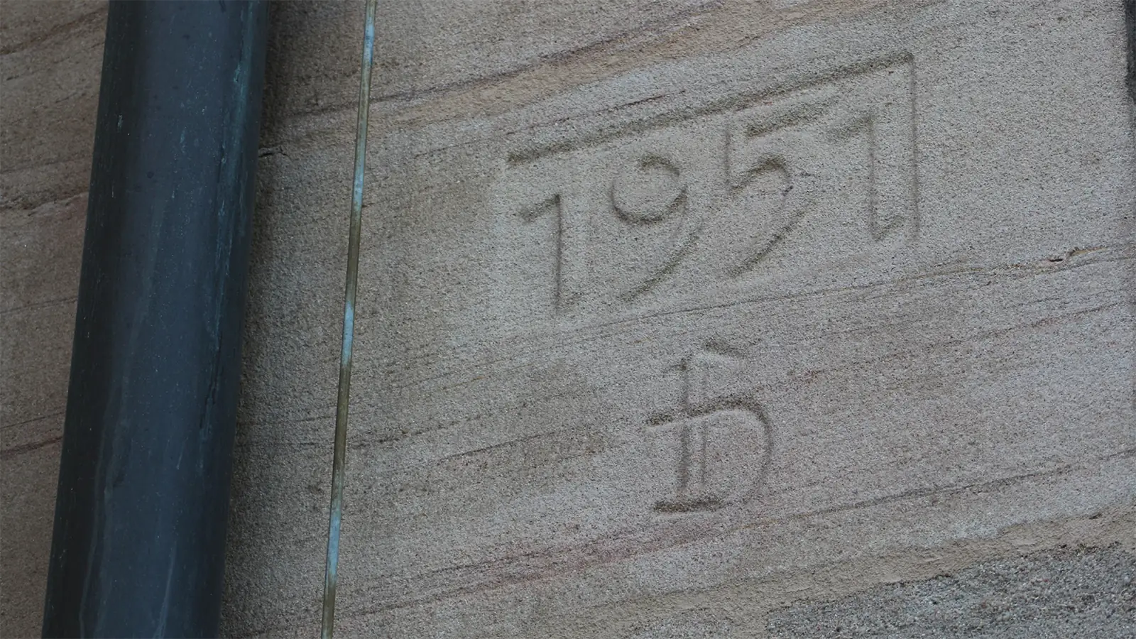 Eine Steinmauer mit der Jahreszahl 1951 und einem eingravierten Schriftzeichens, daneben ein schwarzes senkrechtes Rohr.