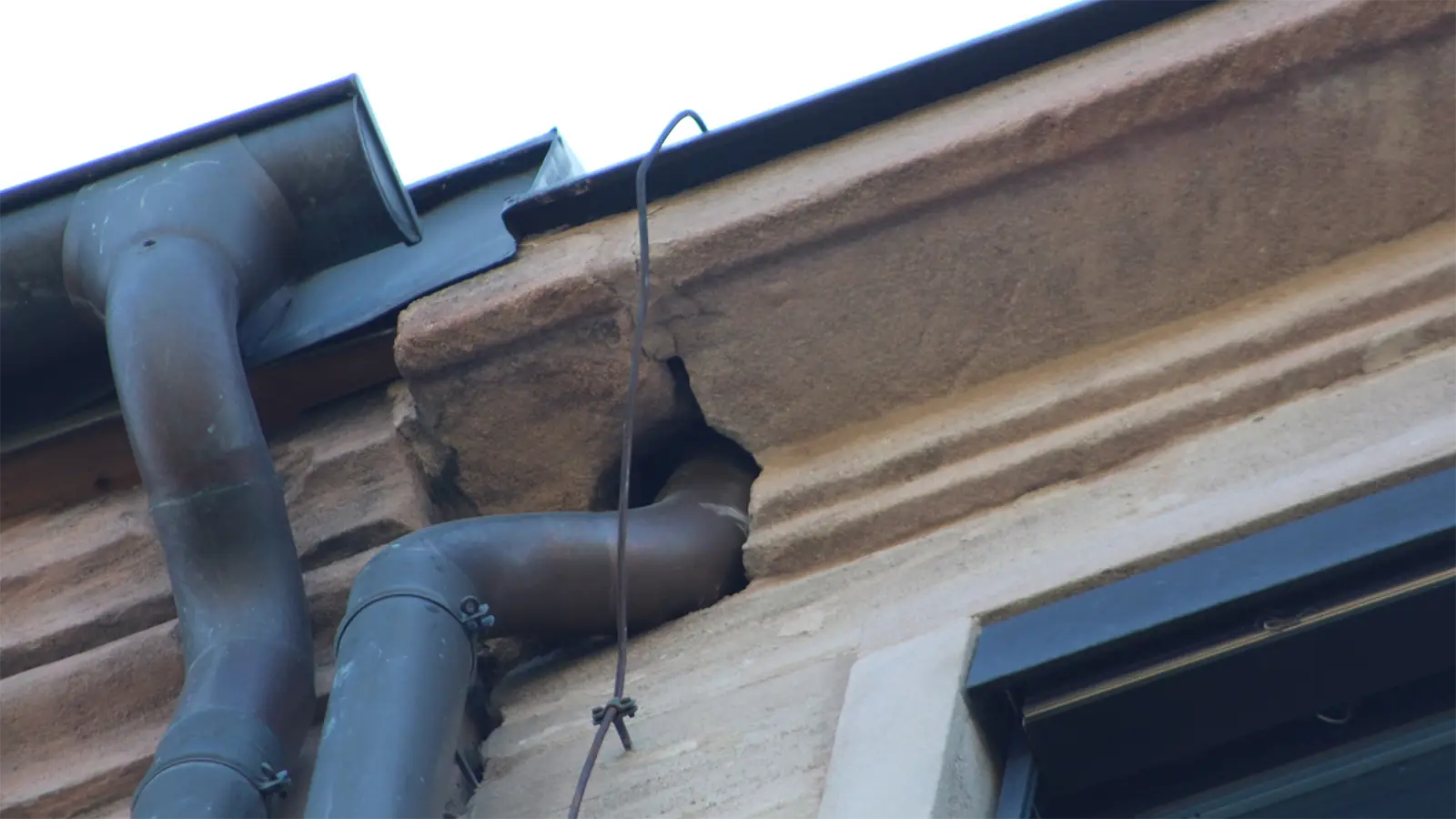 Ein metallenes Abflussrohr mündet in die Seite eines Gebäudes, in dem sich ein großes Loch in der Steinmauer gebildet hat, wahrscheinlich aufgrund von Bauschäden. In der Nähe hängt ein Kabel lose herab.