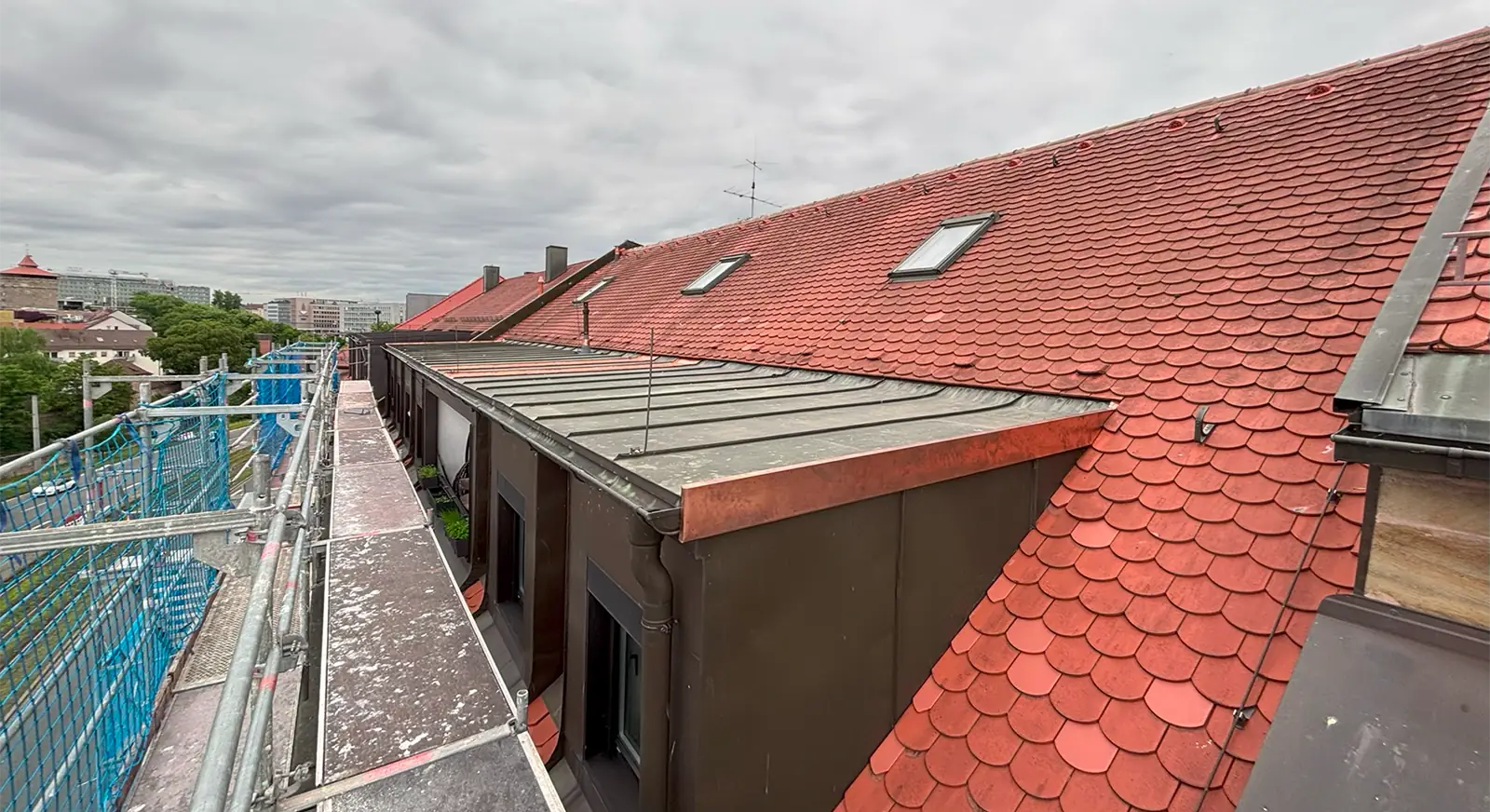 Ein schräges rotes Ziegeldach mit mehreren Oberlichtfenstern, daneben ein mit Metall gedeckter Teil. Ein Baugerüst verläuft entlang der Gebäudekante und bietet einen Blick auf eine Stadt mit Bäumen und bewölktem Himmel.