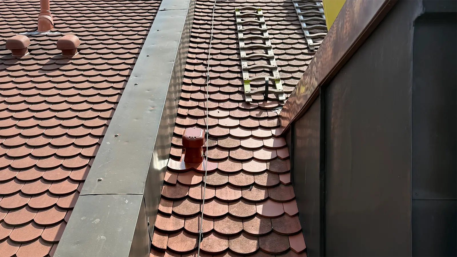 Nahaufnahme eines Ziegeldachs mit sich überlappenden roten Schindeln, Metallvorrichtungen, einer Metallleiter, einem roten Gerät und einem Teil einer dunklen vertikalen Wand auf der rechten Seite. Das Sonnenlicht wirft Schatten auf das Dach.