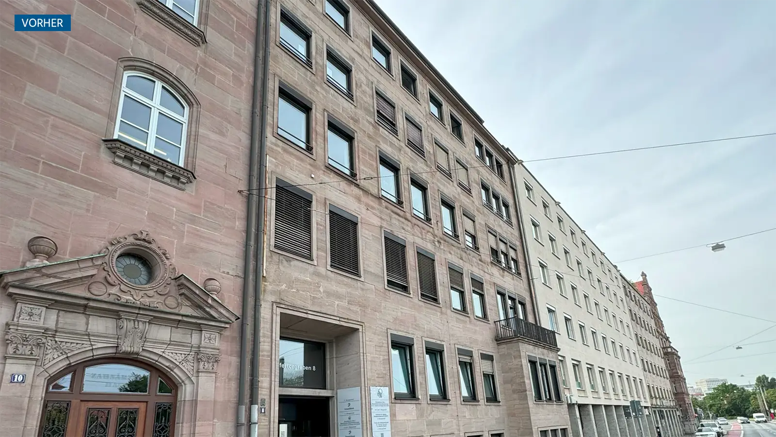 Ein mehrstöckiges Steingebäude mit rechteckigen Fenstern und verzierten architektonischen Details am linken Eingang, gesehen aus einem niedrigen Winkel unter einem bewölkten Himmel.