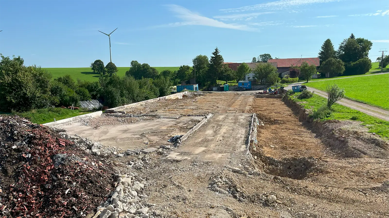 Eine Baustelle mit Dreck- und Schutthaufen liegt neben einer Landstraße, im Hintergrund sind grüne Felder, Bäume, Häuser und eine Windkraftanlage unter einem klaren blauen Himmel zu sehen.