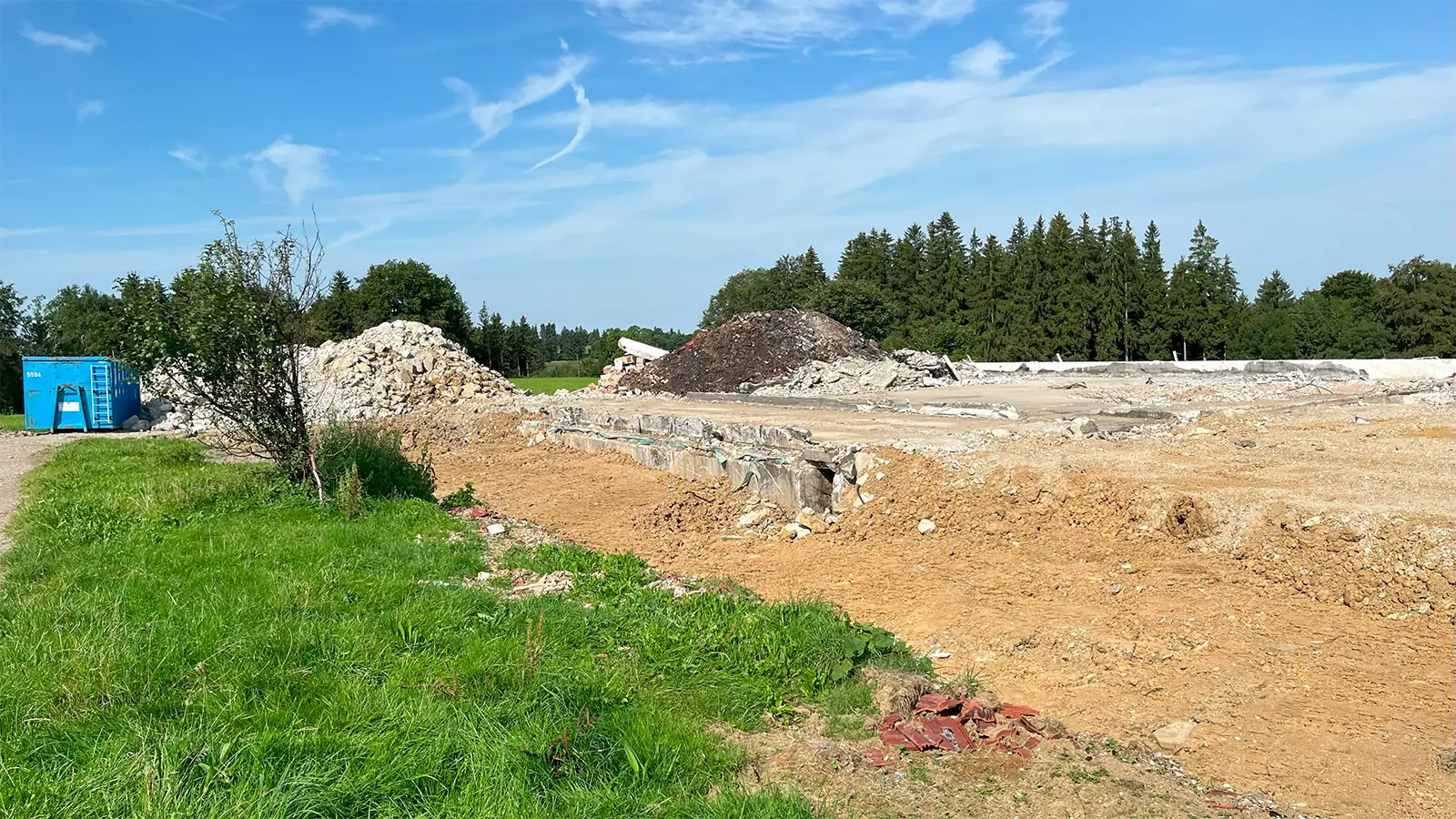 Eine Baustelle mit Schutt- und Erdhaufen, umgeben von Gras und Bäumen unter einem blauen Himmel mit vereinzelten Wolken. Auf der linken Seite des Bildes ist eine blaue Schuttcontainer zu sehen.