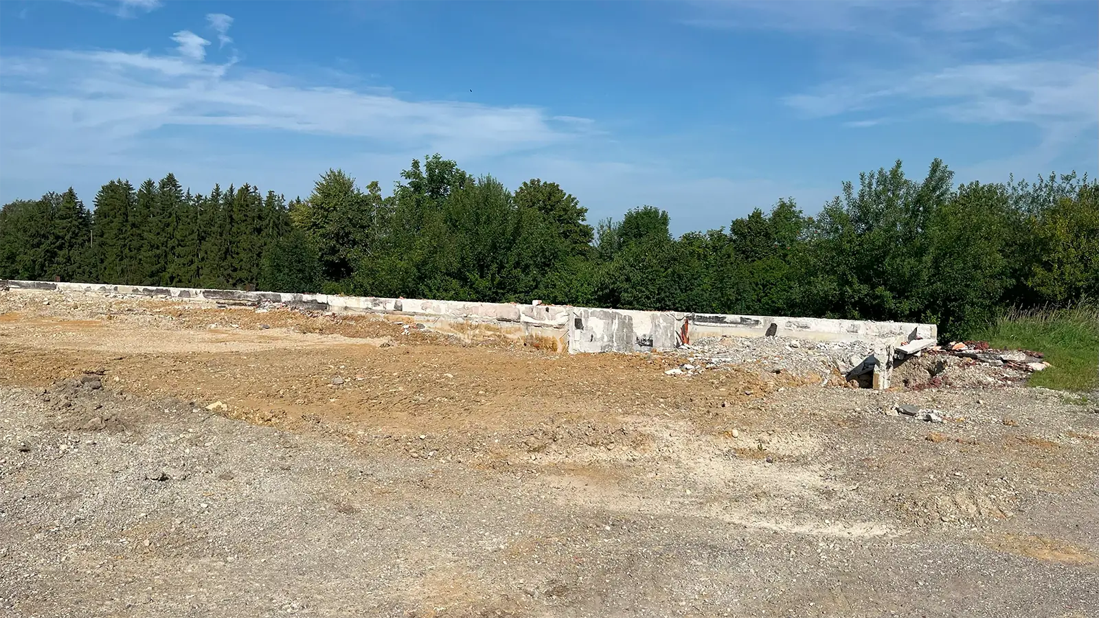 Eine geräumte Baustelle mit nackter, eingeebneter Erde im Vordergrund, die von einer niedrigen Betonmauer begrenzt wird. Grünes Gras und Bäume umgeben das Gelände, und darüber ist ein blauer Himmel mit leichten Wolken zu sehen.