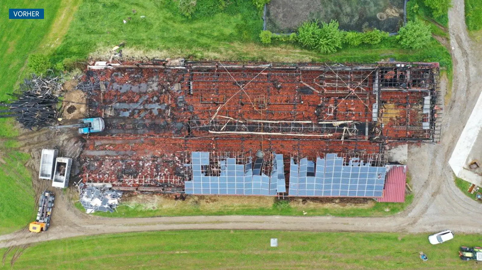 Luftaufnahme eines großen, rechteckigen, verbrannten Gebäudes mit eingestürztem Dach, umgeben von grünem Gras, Feldwegen und Baufahrzeugen. Auf einem Teil des Daches befinden sich noch einige Solarzellen.