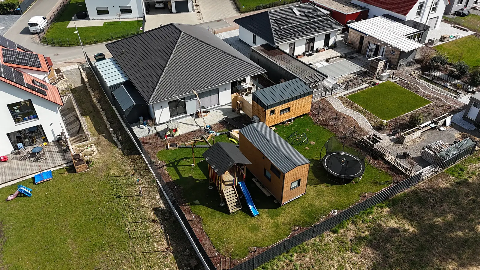 Physiotherpiepraxis aus der Vogelperspektive. Das Gebäude ist einstöckig mit dunklem Dach. Im Gartenbereich wurden temporäre Räume in Modulbauweise bereitgestellt. Um das Gebäude herum sieht man Nachbargrundstücke mit Wohngebäuden.