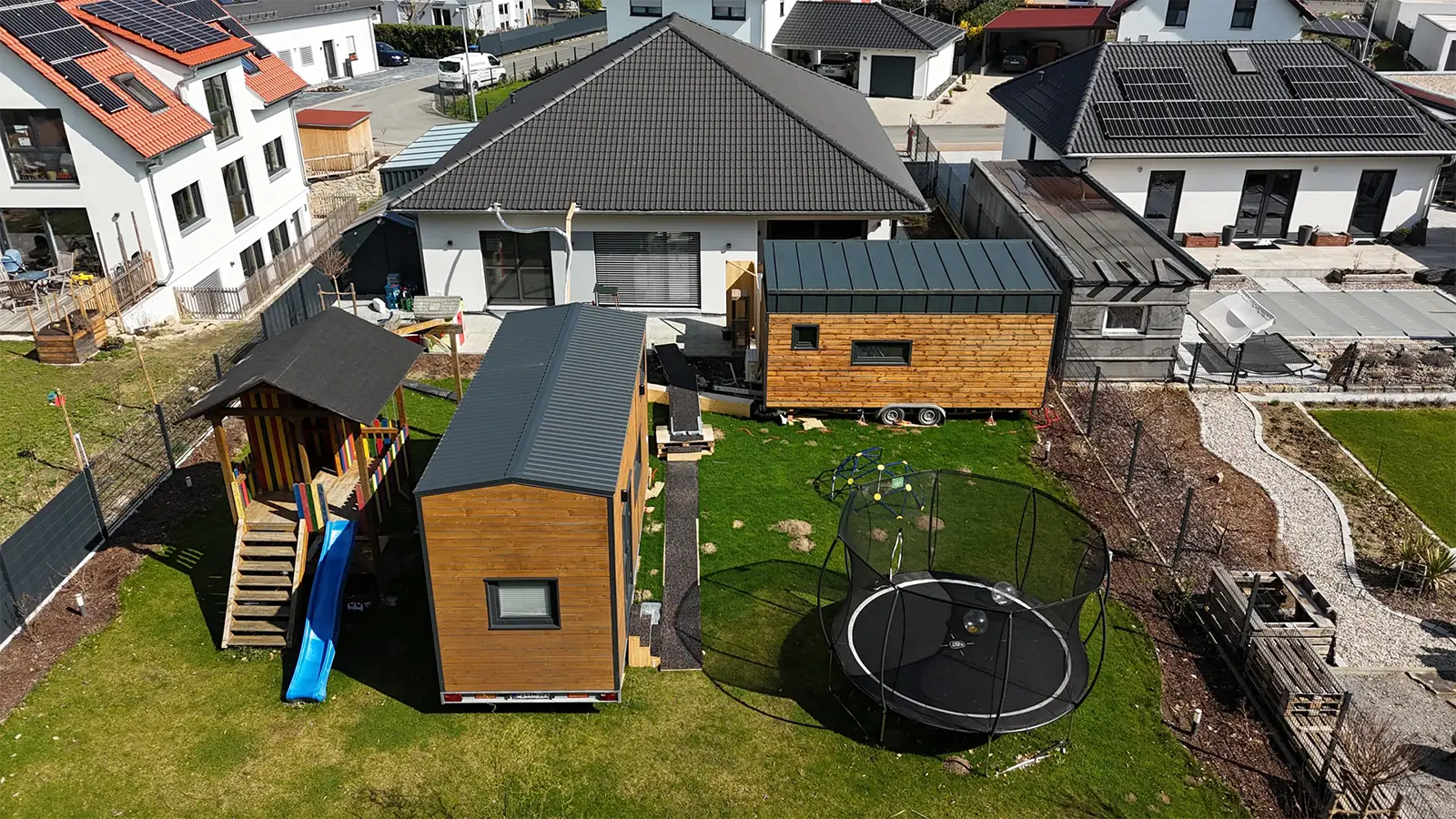 Temporäre Praxisräume in Modulbauweise im Gartenbereich der Praxis. Die Räume sehen aus wie Container mit Wellblechdach und Holzfassade. Neben den bereitgestellten Räumen steht links eine Rutsche mit kleinem Holzhaus für Kinder und rechts ein Trampolin. 