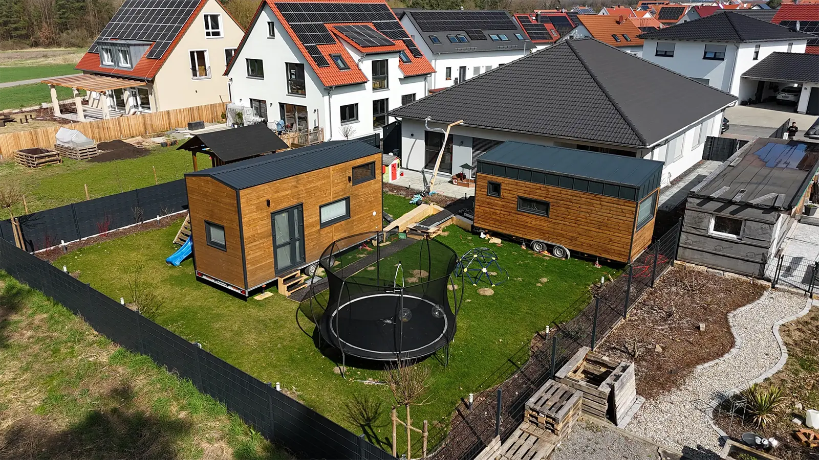 Temporäre Praxisräume in Modulbauweise im Gartenbereich der Praxis. Die Räume sehen aus wie Container mit Wellblechdach und Holzfassade. Neben den bereitgestellten Räumen steht links eine Rutsche mit kleinem Holzhaus für Kinder und rechts ein Trampolin.