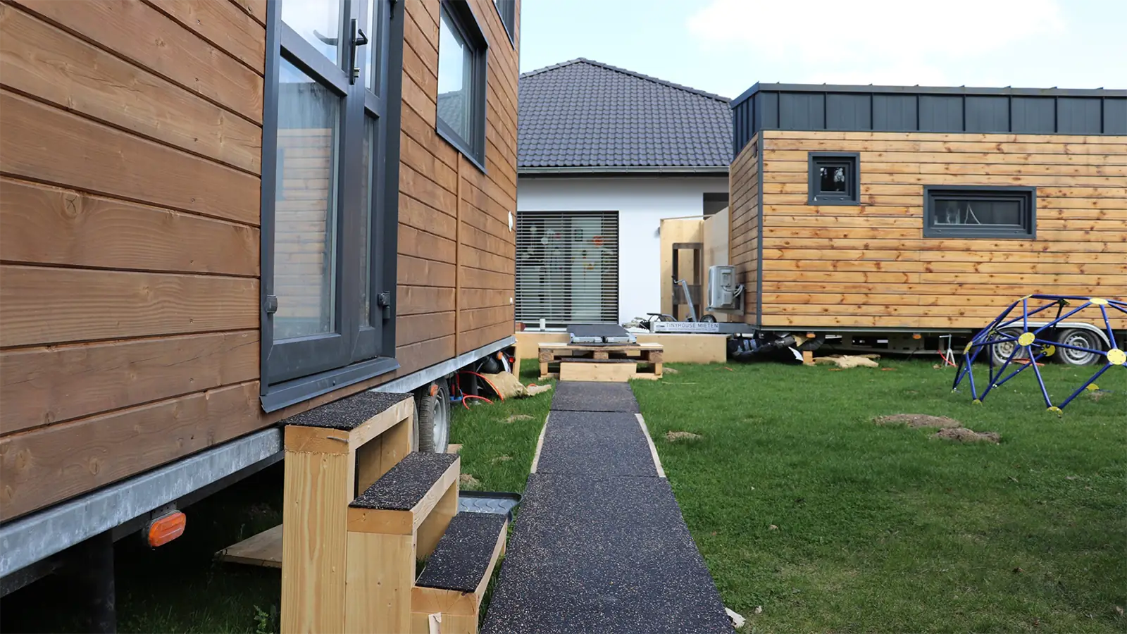 Nahaufnahme der modularen Praxisräume. Die Räume haben eine helle Holzfassade und werden durch künstlich angelegte Wege verbunden. Im Vordergrund führt eine Holztreppe zur Tür einer der Räume.