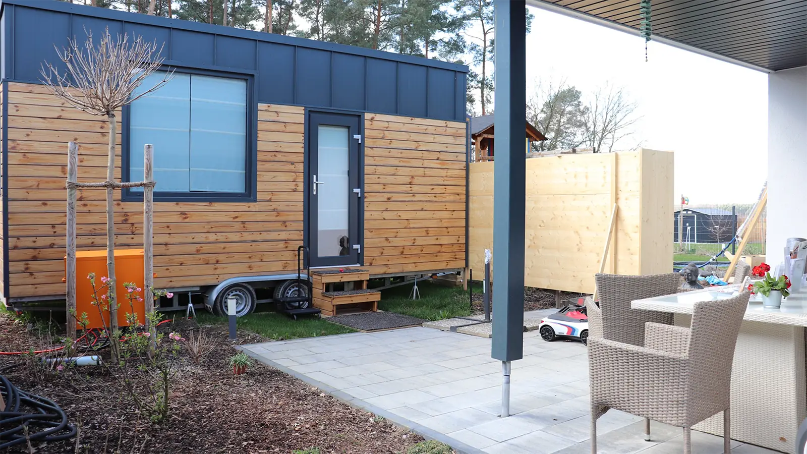 Modularer Praxisraum mit heller Holzfassade. Eine kleine Holztreppe führt zur Tür des Raumes. Im Vordergrund ist die Terrasse der Praxis und Korbstühle zu erkennen.