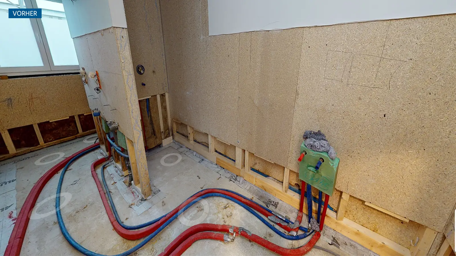 Innenansicht der Praxis während der Sanierung. Die Schäden wurden beseitigt. Wände und Boden sind unverkleidet. Auf dem Boden liegen Rohre und Kabel, die zu einem Wandanschluss führen.