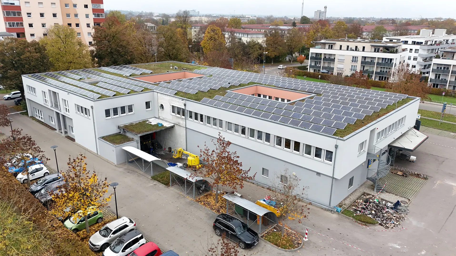 Supermarkt aus der Vogelperspektive. Das Gebäude ist rechteckig mit begrüntem Flachdach. Auf dem Dach befinden sich auf voller Länge Solarzellen. Um das Gebäude sind Fahrwege, Beladungszone und Parkplätze. Im Hintergrund sieht man Wohngebäude.