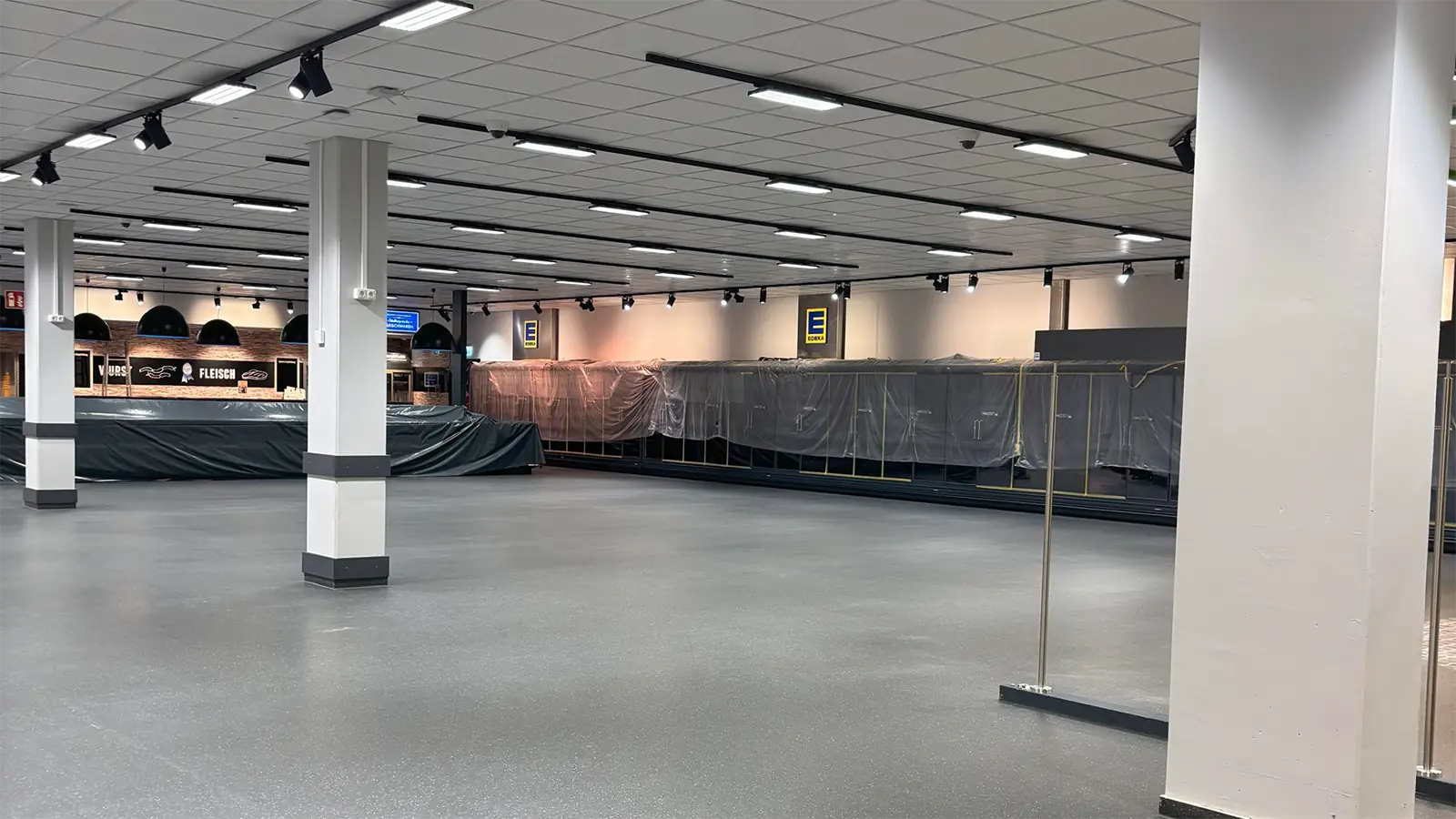 Leere Verkaufsfläche des Supermarkts nach der Sanierung. Im Hintergrund sind abgedeckte Kühltruhen und Regale zu erkennen. An den Wänden ist das Logo des Unternehmens angebracht. Der Raum ist sauber und ordentlich.
