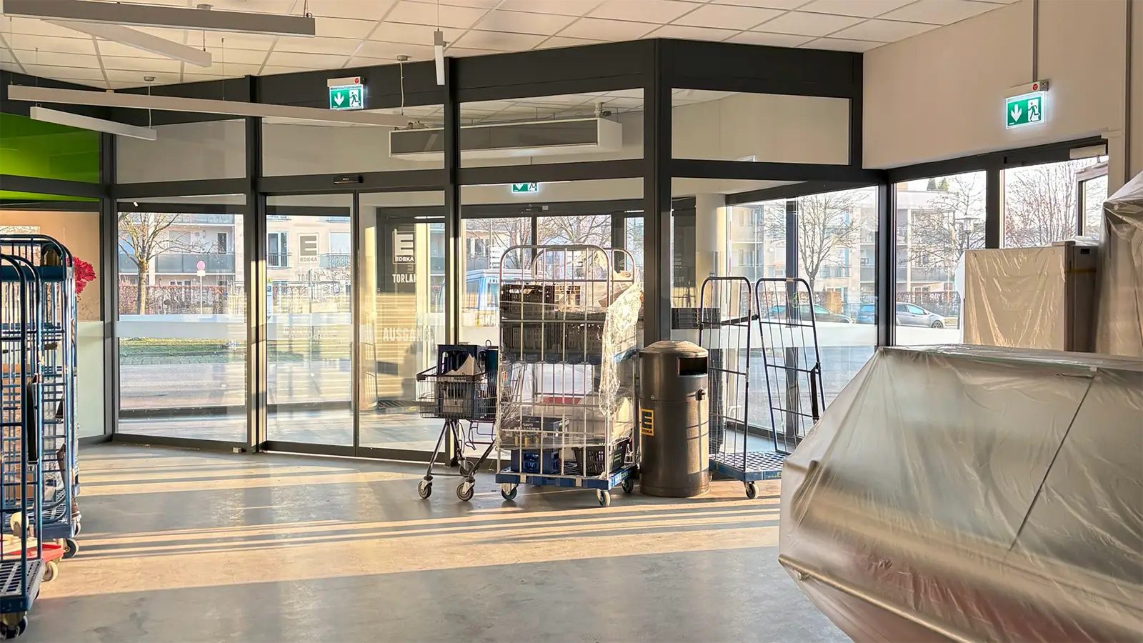 Eingangsbereich des Supermarkts nach der Sanierung. Im Vordergrund stehen abdeckte Kühltruhen und Warentransportwägen. Im Hintergrund sind die gläsernen Schiebetüren zu erkennen.