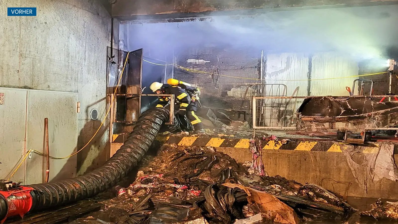 Brandschaden im Anlieferungsbereich des Supermarkts. Feuerwehrkräfte arbeiten mit einem großen Rohr an der Beseitigung von Feuer und Rauch. Rauch durchzieht den Raum. Im Vordergrund liegt verbrannter Schutt.