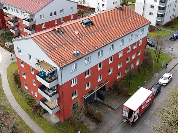 Vogelperspektive eines Mehrfamiliengebäudes. Das Gebäude hat drei Stockwerke. Bis zum zweiten Stock ist die Fassade rot verputzt, im dritten Stockwerk ist die Fassade grau. Um das Gebäude stehen weitere Mehrfamilienhäuser und Grünflächen.