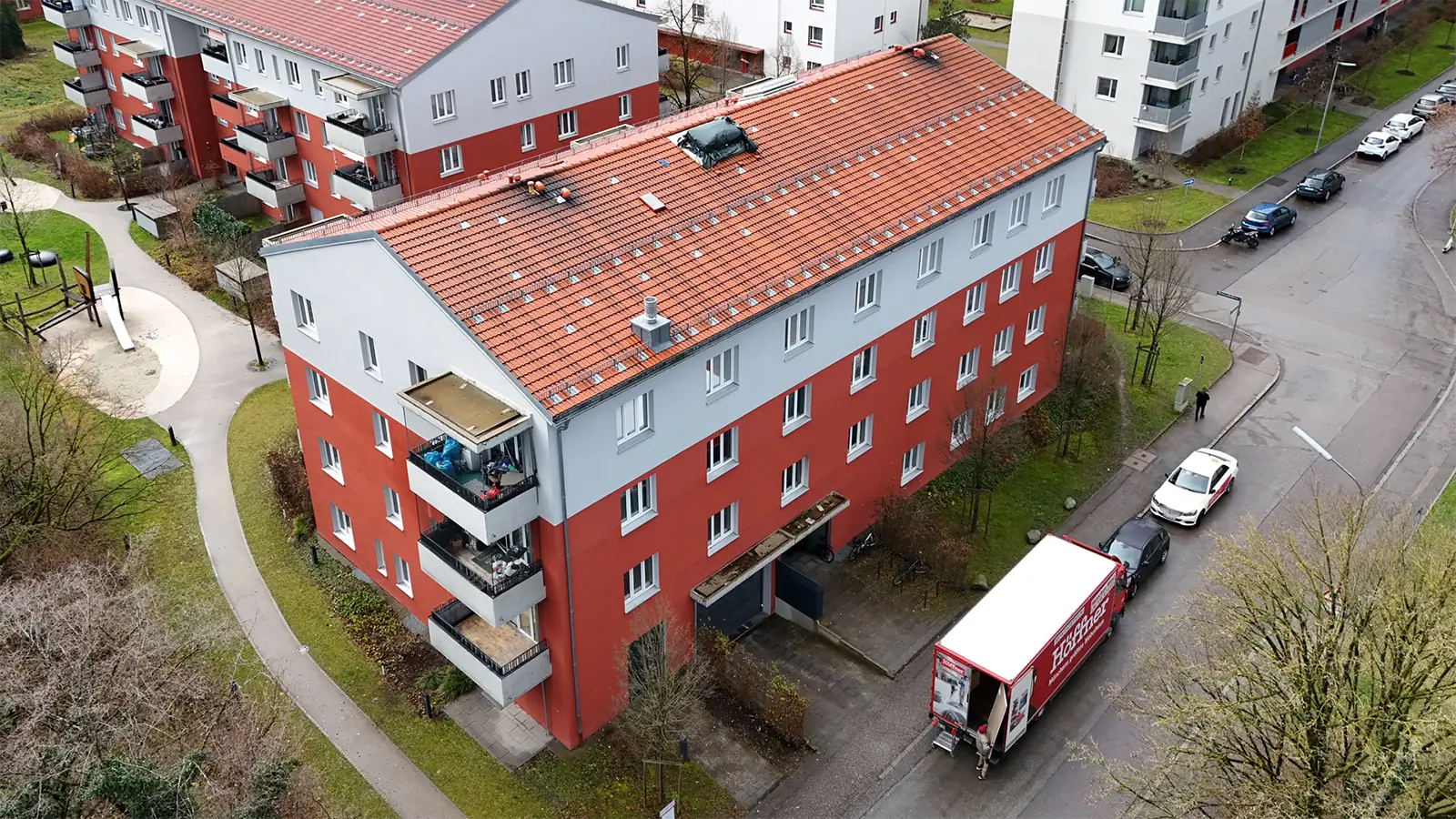Vogelperspektive eines Mehrfamiliengebäudes. Das Gebäude hat drei Stockwerke. Bis zum zweiten Stock ist die Fassade rot verputzt, im dritten Stockwerk ist die Fassade grau. Um das Gebäude stehen weitere Mehrfamilienhäuser und Grünflächen.