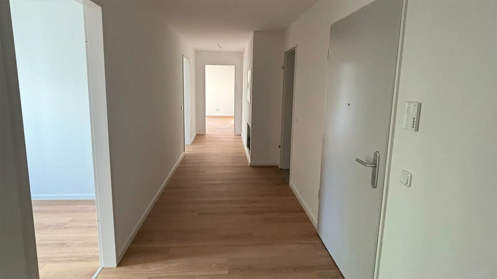 Flur in der renovierten Wohnung. Die Wände und Decke sind weiß gestrichen, der Fußboden ist in heller Holzoptik. Im Flur sind fünf Durchgänge und Türen zu sehen, einer davon ist die geschlossene Wohnungstür.