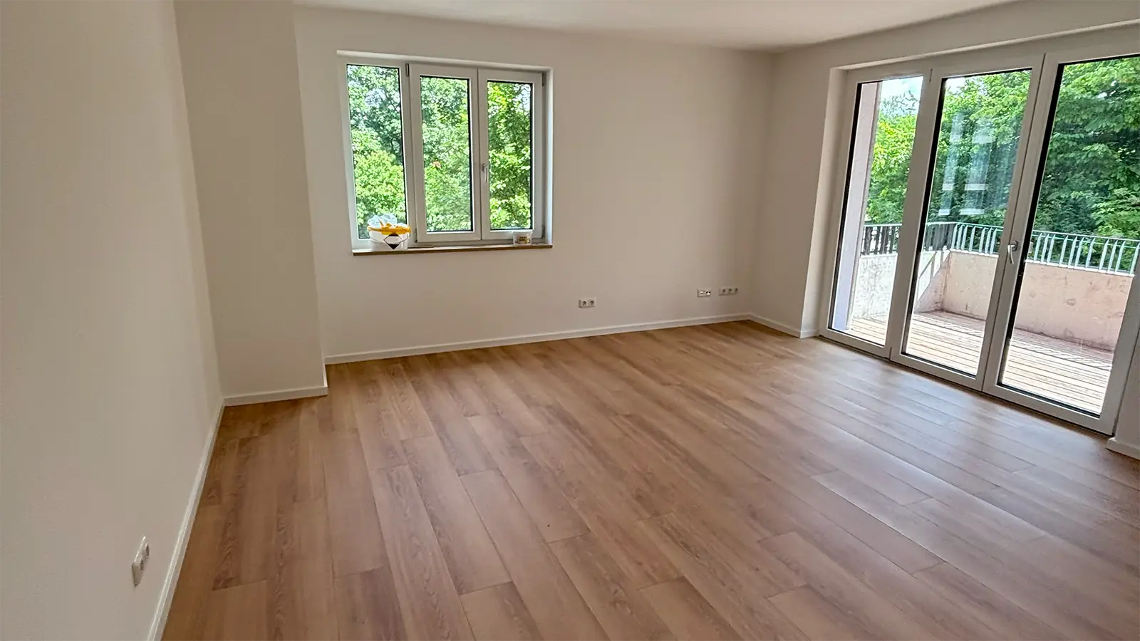 Raum in der renovierten Wohnung. Wände und Decke sind weiß gestrichen, der Fußboden ist in heller Holzoptik. Durch ein Fenster an der hinteren linken Wand sind Laubbäume zu erkennen. An der rechten Wand führt eine Glastüre auf den Balkon.