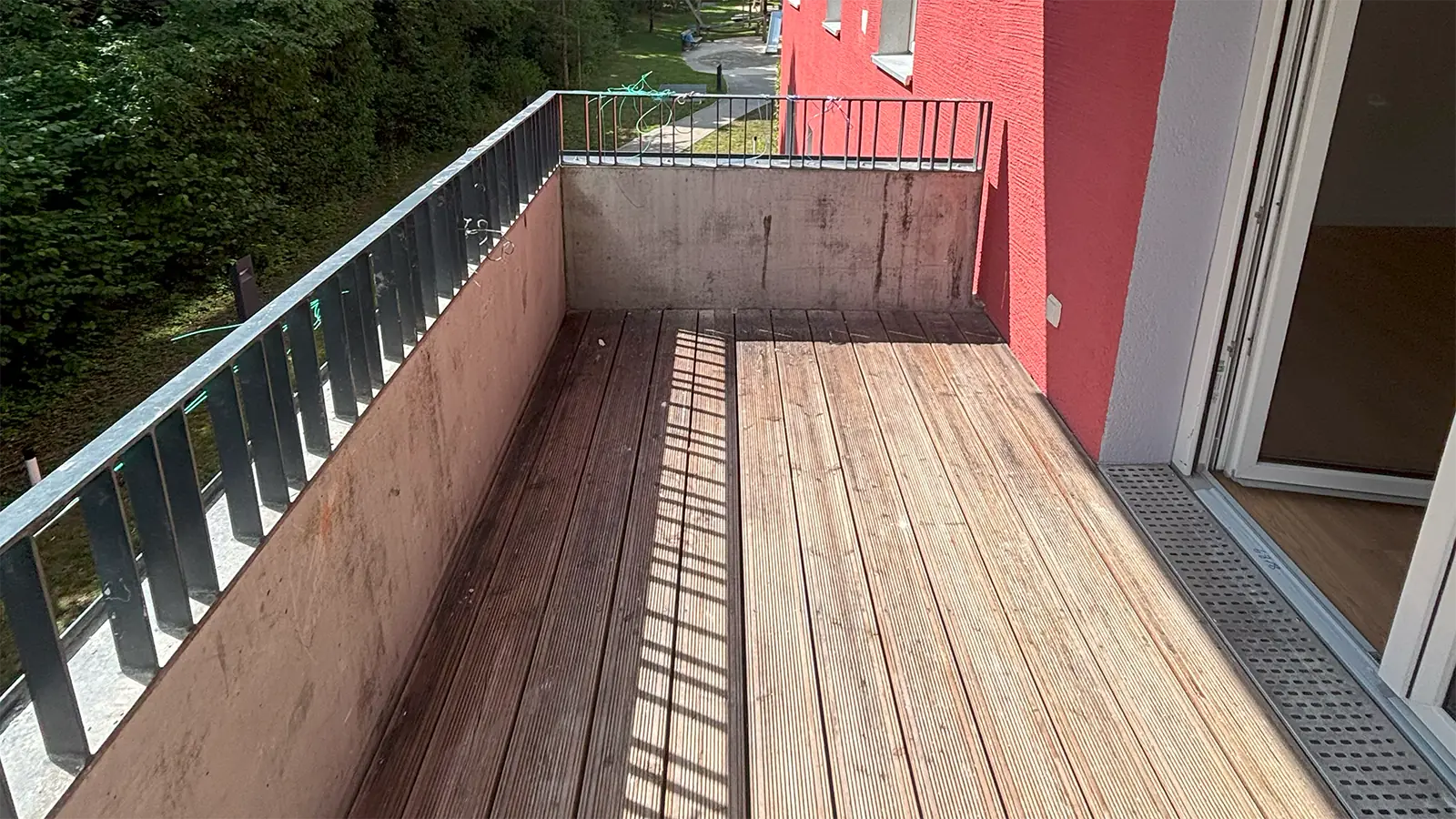 Balkon der renovierten Wohnung mit Blick in die Natur. Das Geländer ist hüfthoch aus Beton und Stahl. Die Außenwand trägt eine rote Putz-Fassade. Der Boden des Balkons ist in Holzoptik.