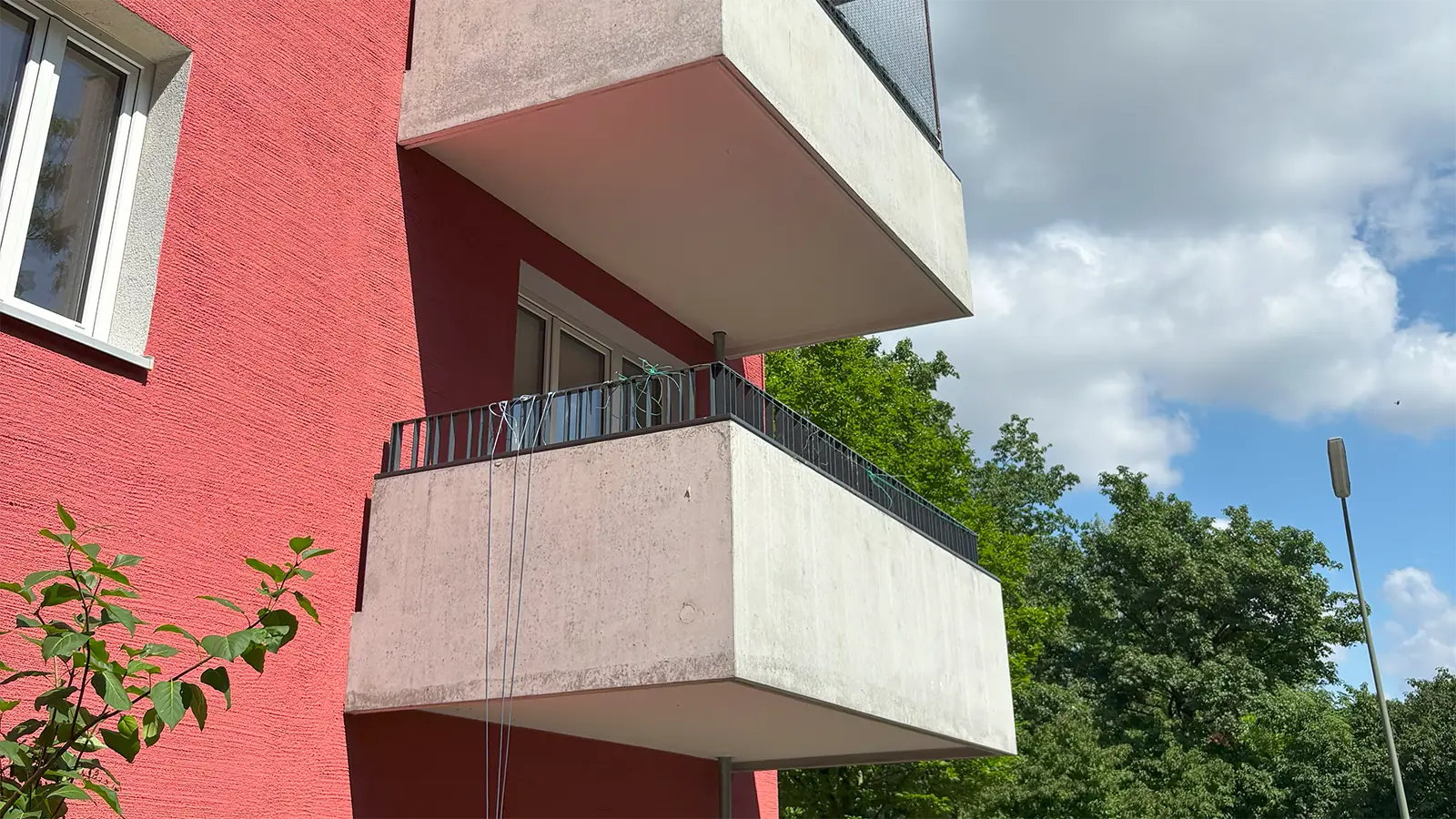 Außenansicht auf den Balkon der renovierten Wohnung. Im Vordergrund ist die rote Fassade des Hauses zu sehen, über dem Balkon sieht man den Balkon der darüberliegenden Wohnung. Im Hintergrund sind Bäume und eine Straßenlaterne zu erkennen.