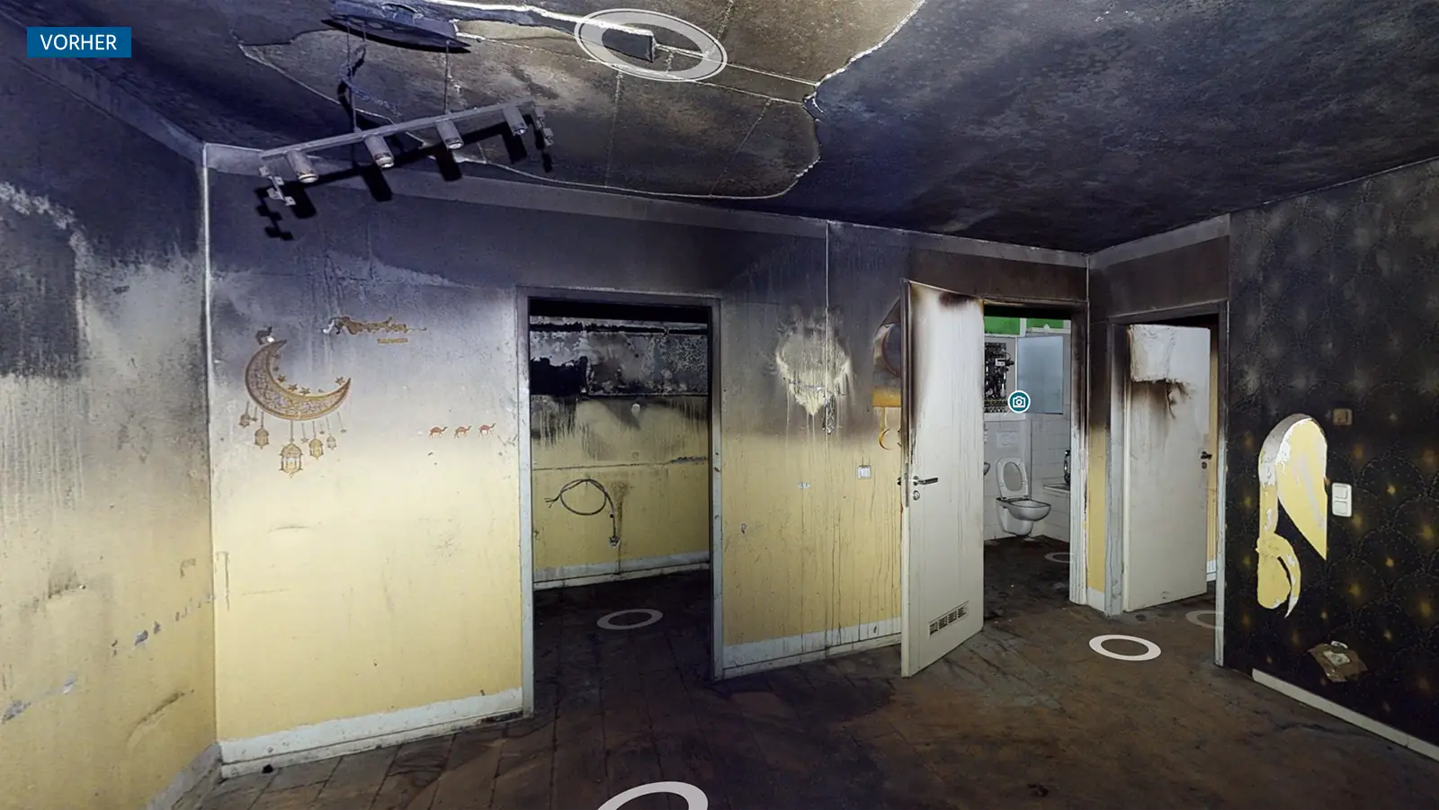 Brandschaden im Raum einer Wohnung. Wände und Decke sind verrußt. Der Putz in der Decke hat ein großes Loch. Durch drei offene Türen kann in andere, verrußte Räume geblickt werden. Der Boden ist voller Asche und Dreck.