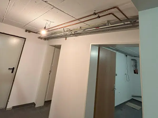 Kellergeschoss eines Wohngebäudes nach der Sanierung. Drei Türen führen zu anliegenden Räumen. An der Decke sind Rohre zu erkennen. Eine Glühbirne erleuchtet den Raum.