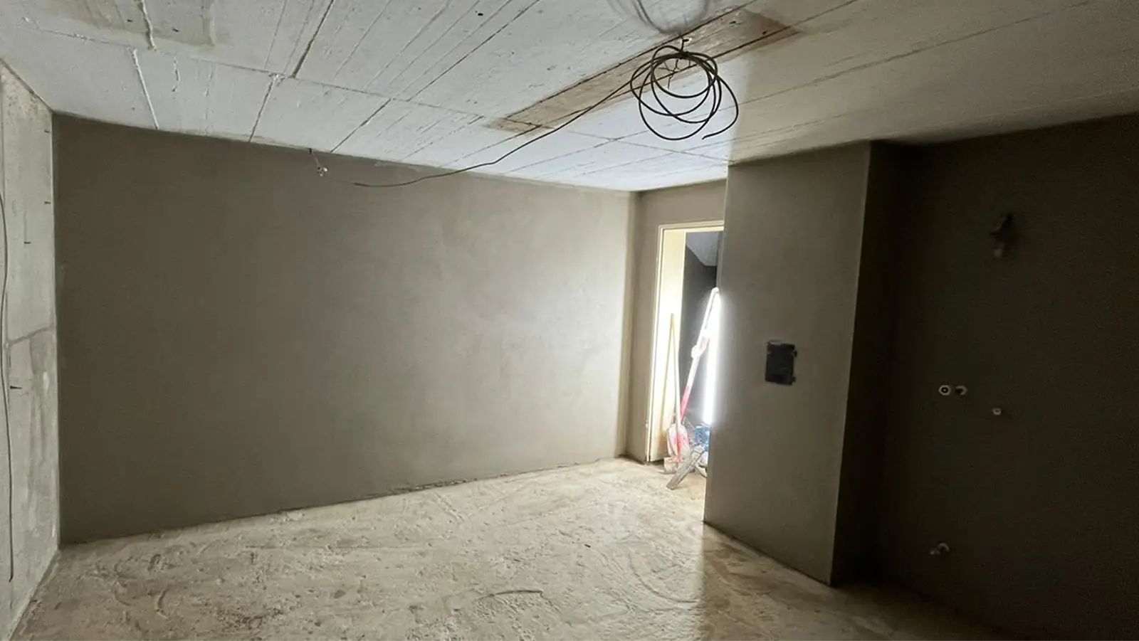 Ein Kellerraum während der Sanierung. Boden, Wand und Decke sind noch unbehandelt und unverputzt. Im Hintergrund ist eine offene Tür zu sehen, aus der ein Licht in den Raum scheint. An der Decke sind offene Kabel zusammengebunden.