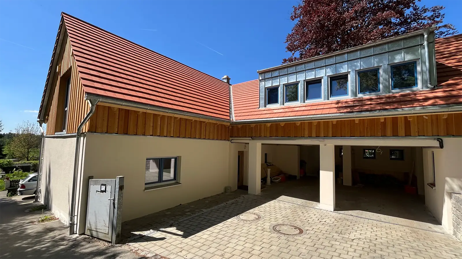 Fertig saniertes Haus nach Brandschaden. Das Dach ist neu gedeckt, die Fassade ist erneuert. Das obere Dachgeschoss trägt eine Holzfassade, das Ergeschoss wurde in hellem Gelb verputzt. Die Garagen sind offen und bieten einen Blick ins saubere Innere.