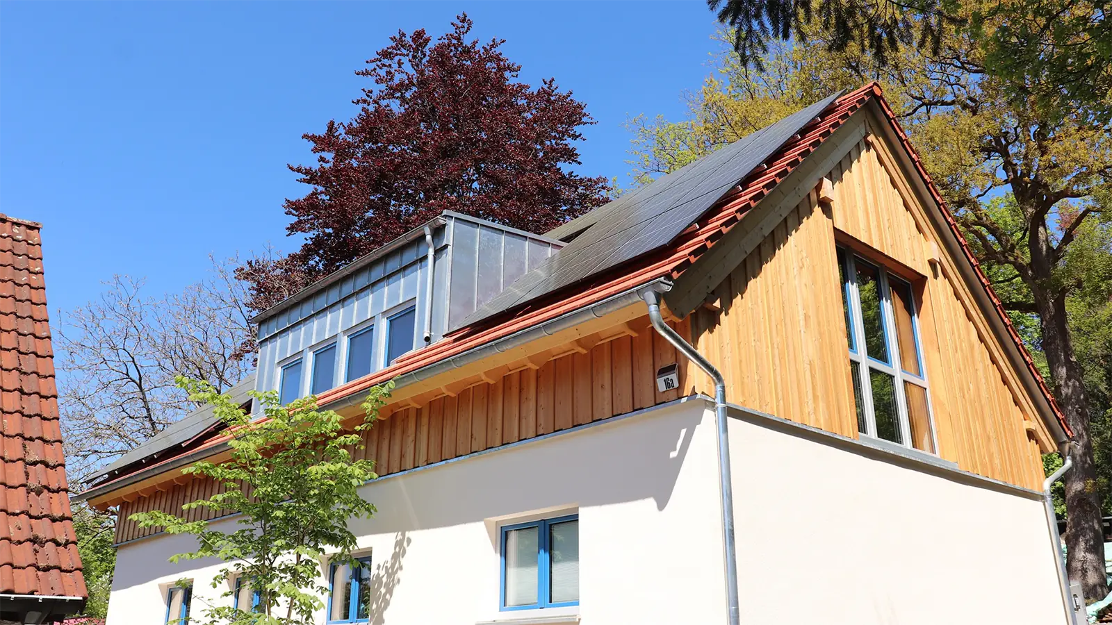 Fertig saniertes Haus nach Brandschaden. Die Fassade des Dachgeschosses ist aus Holzlatten, die Fassade des Erdgeschosses wurde in einem gelblichen Weiß verputzt. Auf dem Dach sind Solarzellen angebracht.