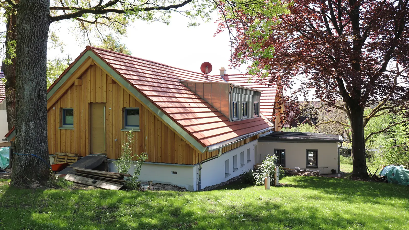 Rückansicht des fertig sanierten Hauses. Im Vordergrund ist ein Zugang zum Dachgeschoss von einem grasigen Hügel zu erkennen. Das Dach wurde mit roten Ziegeln gedeckt. Um das Haus sind Bäume und Grünflächen zu sehen.